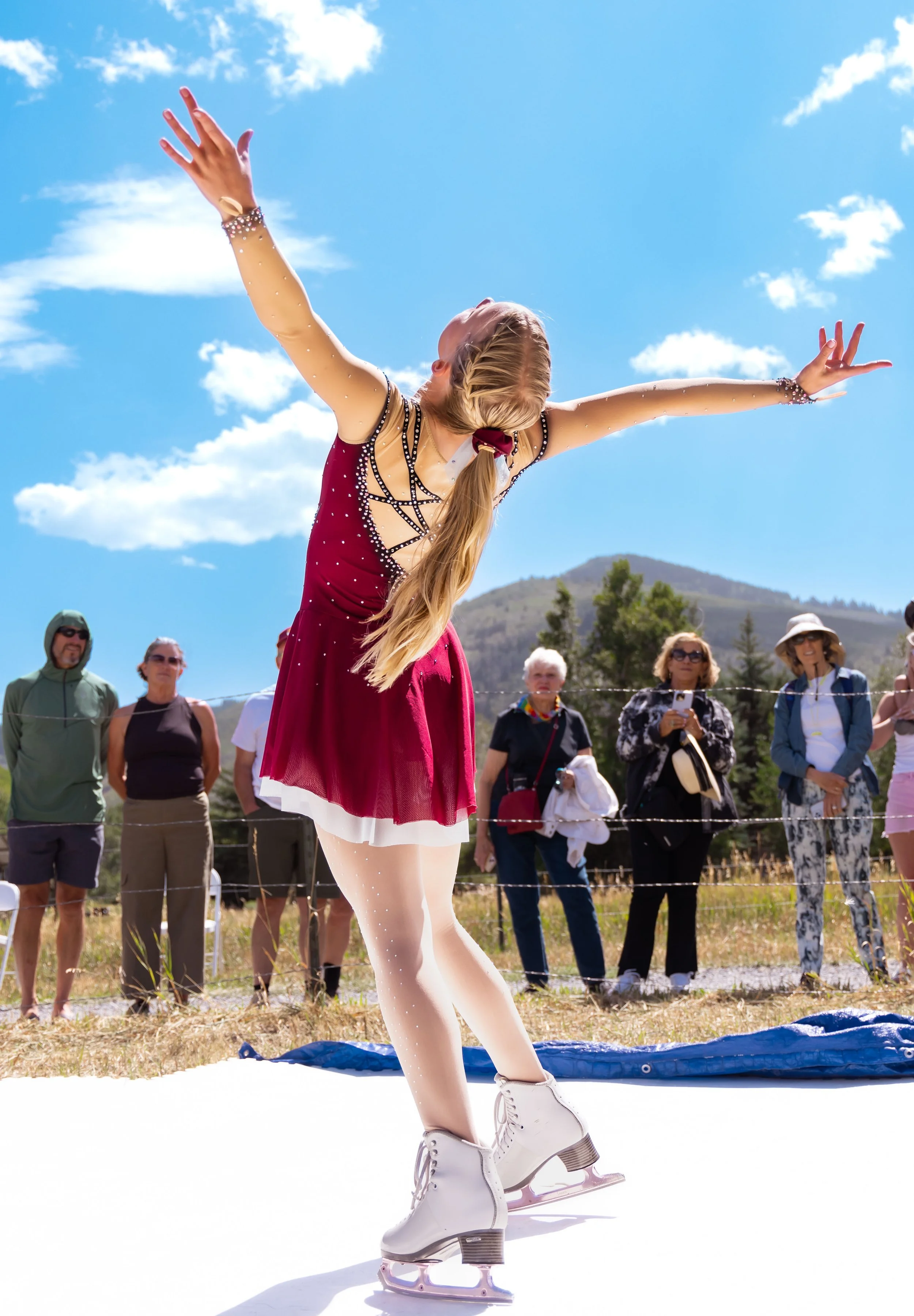 A young woman figure skates outdoors on a sunny day, with arms extended and head tilted back, while spectators watch behind a barrier in a scenic mountainous area.