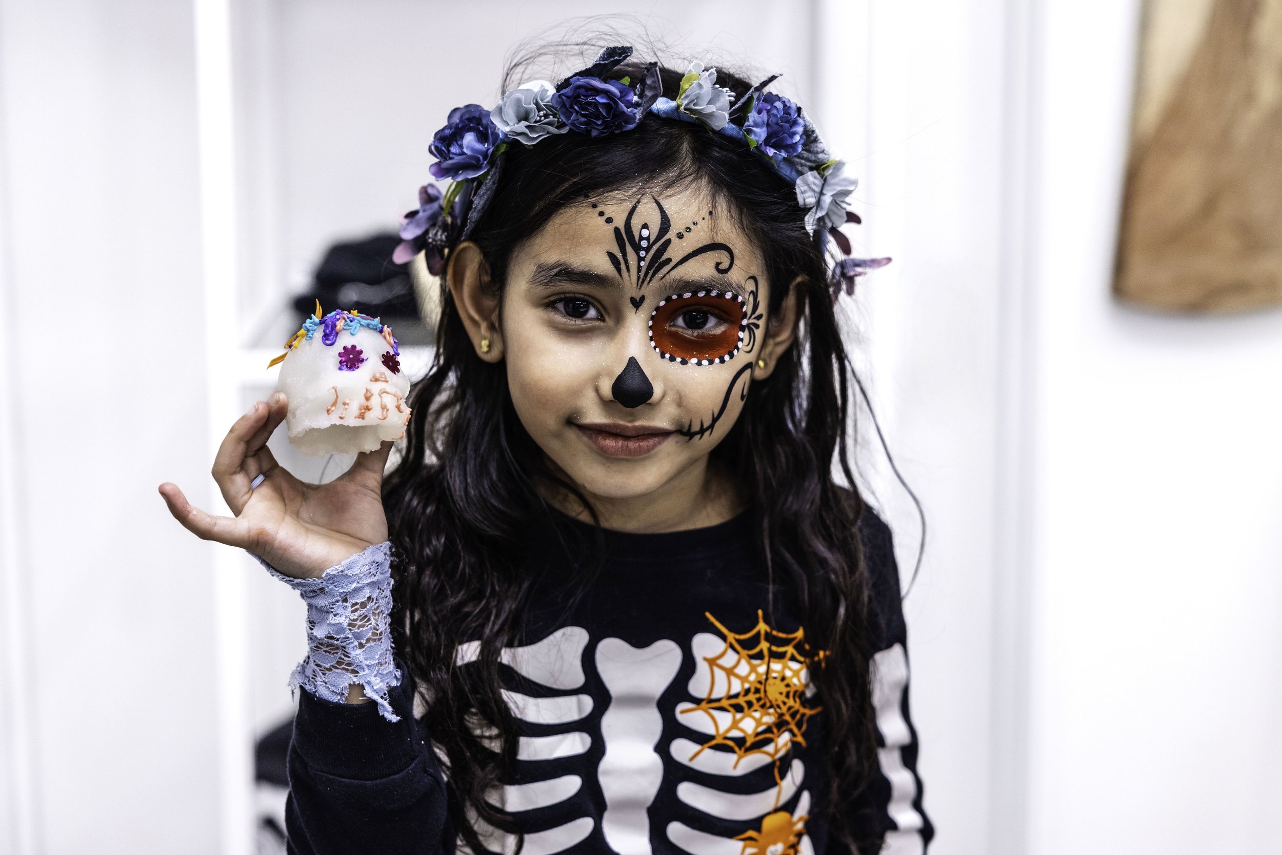 Young girl with face paint in Día de los Muertos style, wearing a skull skeleton shirt, holding a decorated sugar skull candle, and wearing a blue floral crown.