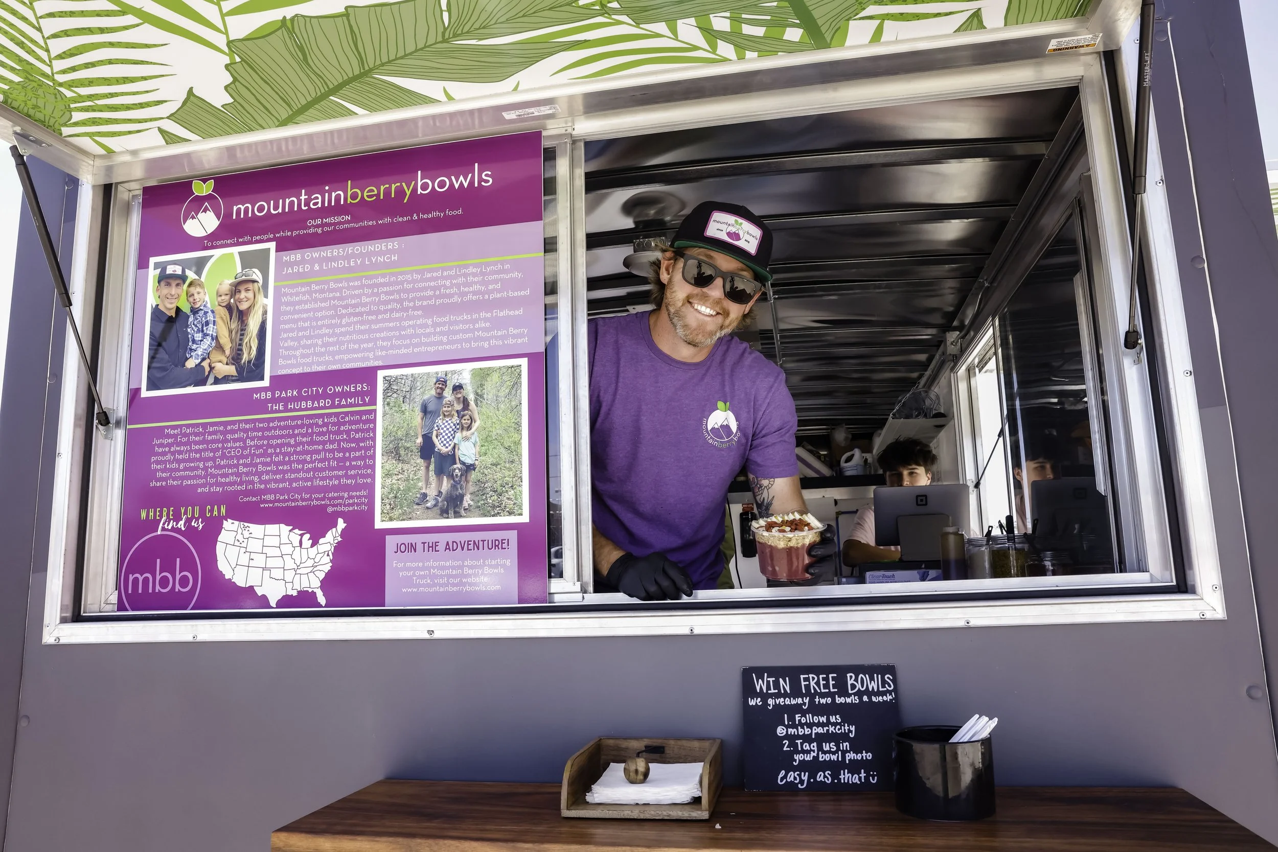 A man with sunglasses, a purple t-shirt, and a cap is smiling and holding a bowl of ice cream or frozen yogurt in a food truck. There are two other people in the background inside the truck.