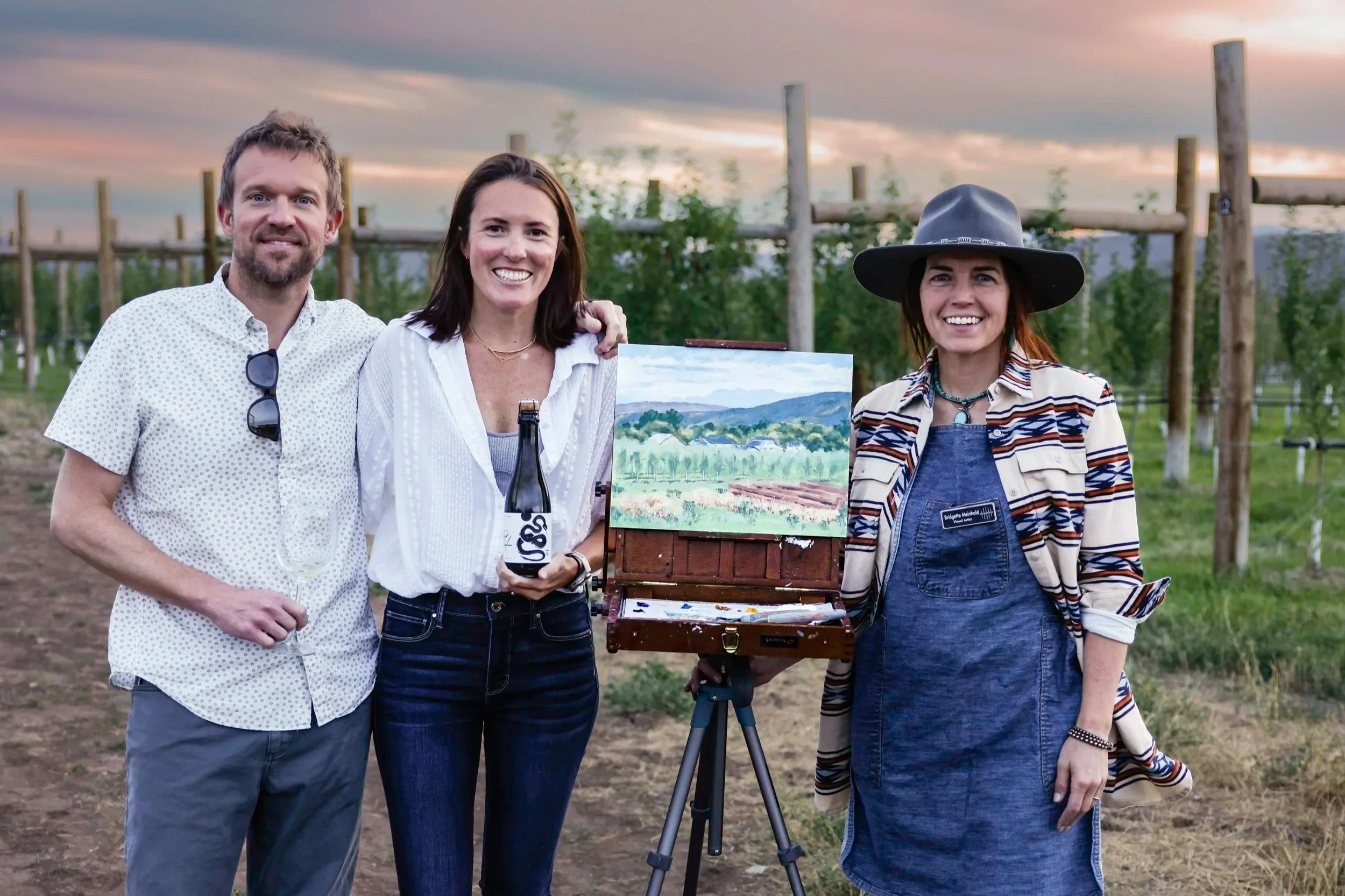 Three people standing outdoors during sunset with an artist's easel displaying a landscape painting, surrounded by greenery and farm structures in the background.