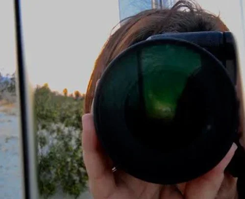 Person taking a photo with a camera, reflected in a mirror, outdoors with trees in the background.