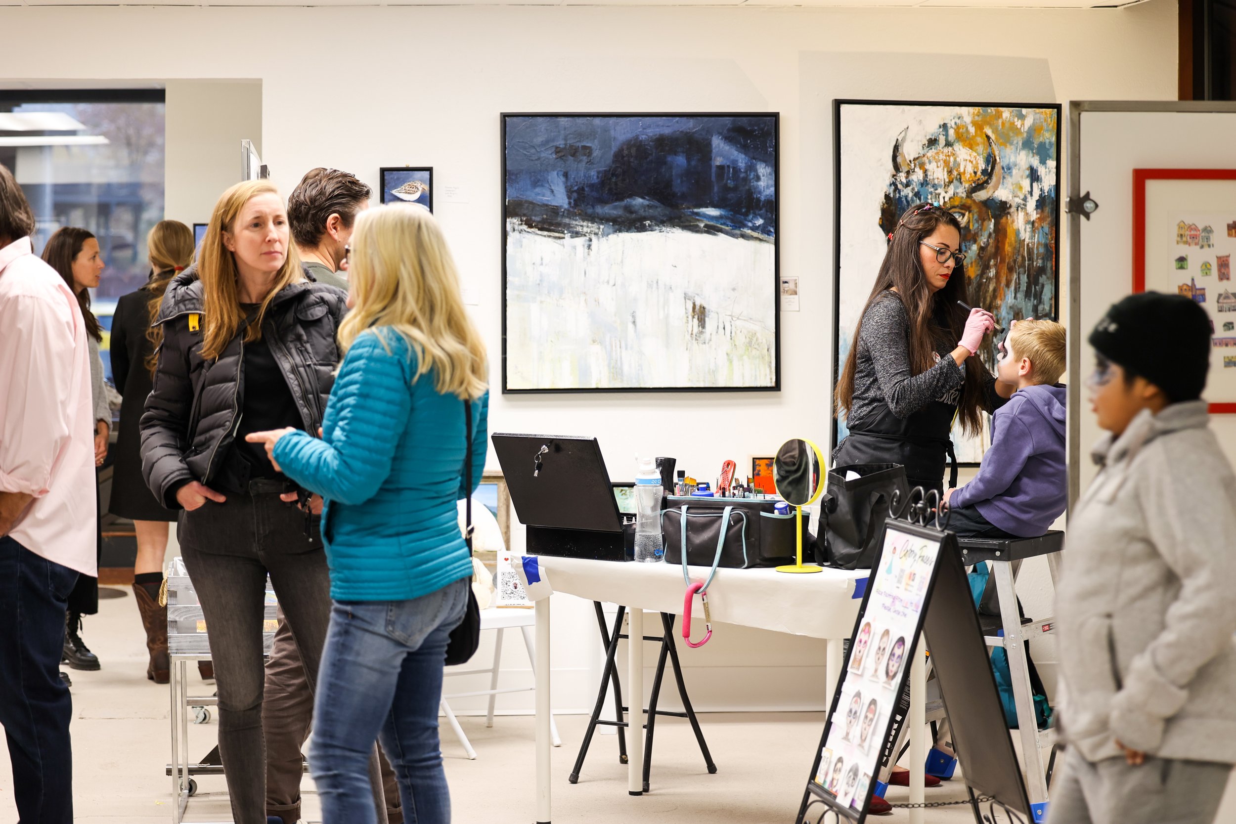 Pessoas numa galeria de arte, com duas mulheres a conversar em primeiro plano, uma menina a pintar o rosto e outros visitantes a ver pinturas nas paredes.