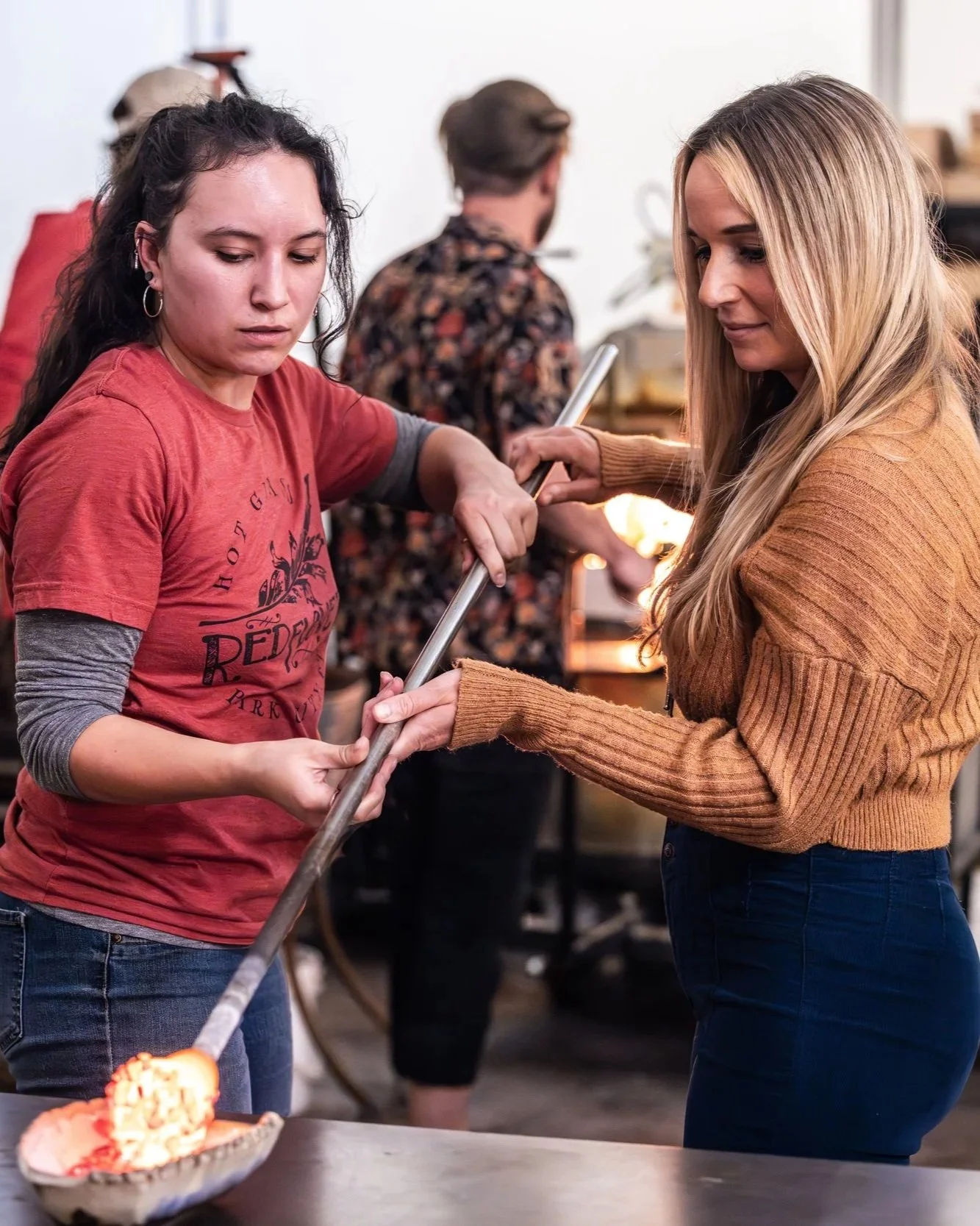 A woman in a brown sweater and another woman in a red t-shirt are holding a long metal tool together, making blown glass art. They are inside a room, with a man in a floral shirt in the background.