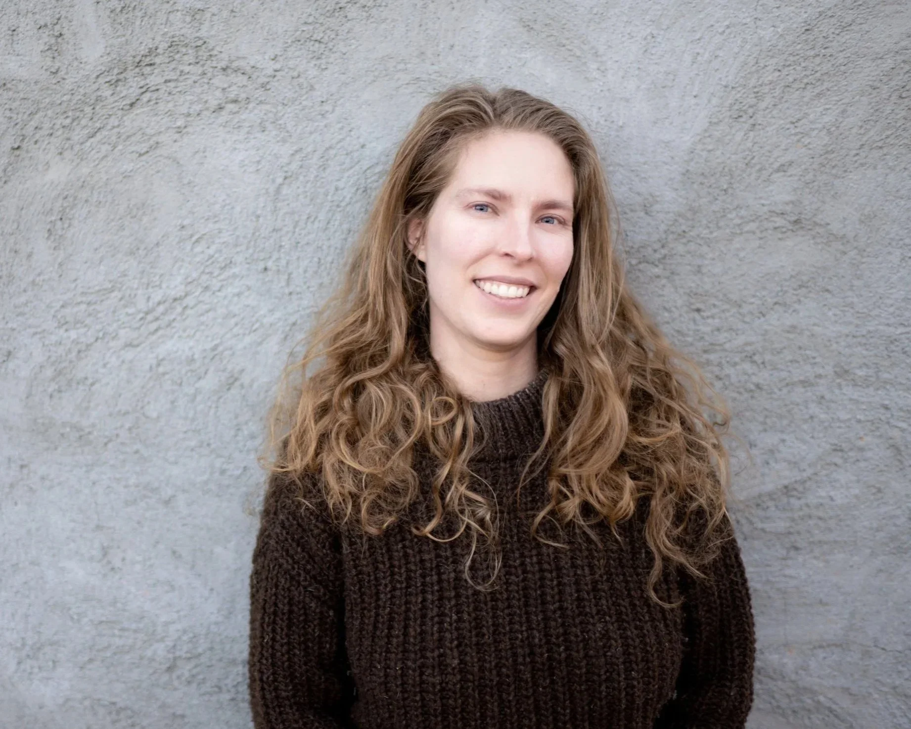 A young woman with long, curly brown hair, smiling, standing against a textured gray wall, wearing a dark brown sweater.