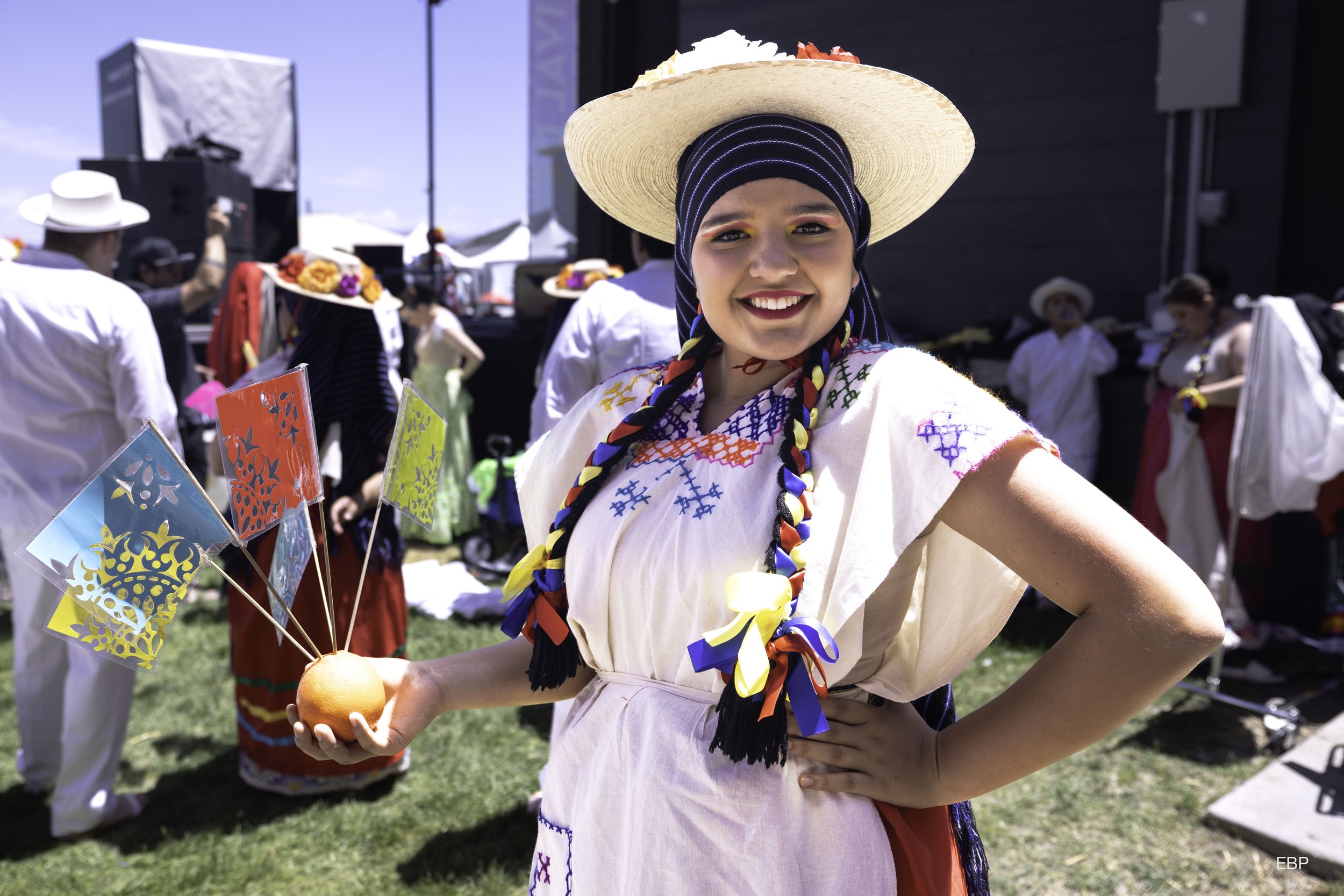 A young woman smiling at a cultural festival, wearing traditional Latino folk attire with a wide-brimmed straw hat and braided ribbons, holding a small orange with colorful paper flags.