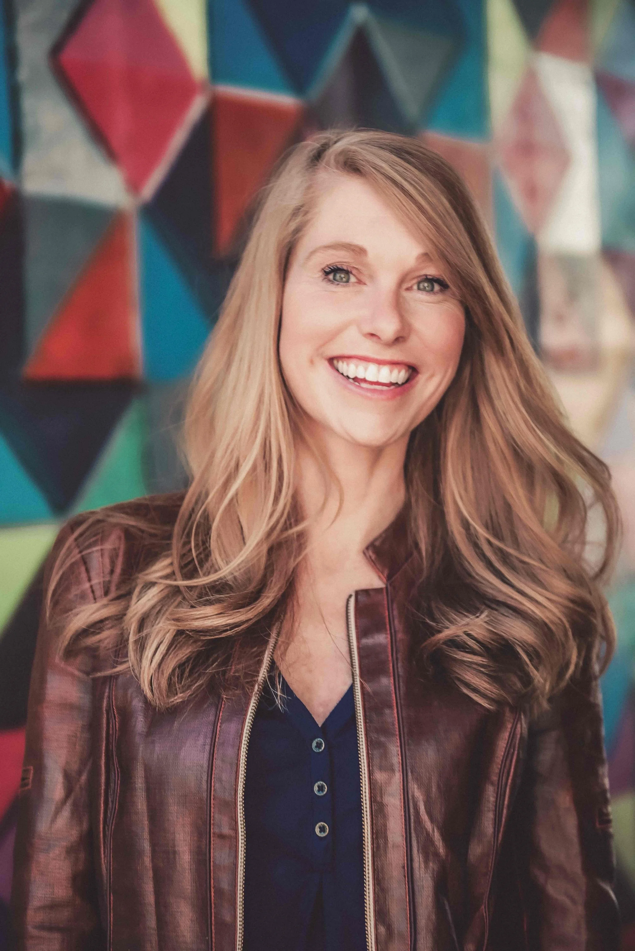 A woman with long, wavy red hair smiling in front of a colorful geometric mural.