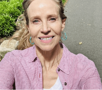A woman smiling outdoors, wearing a pink and white striped shirt and hoop earrings, standing on a paved path with greenery nearby.