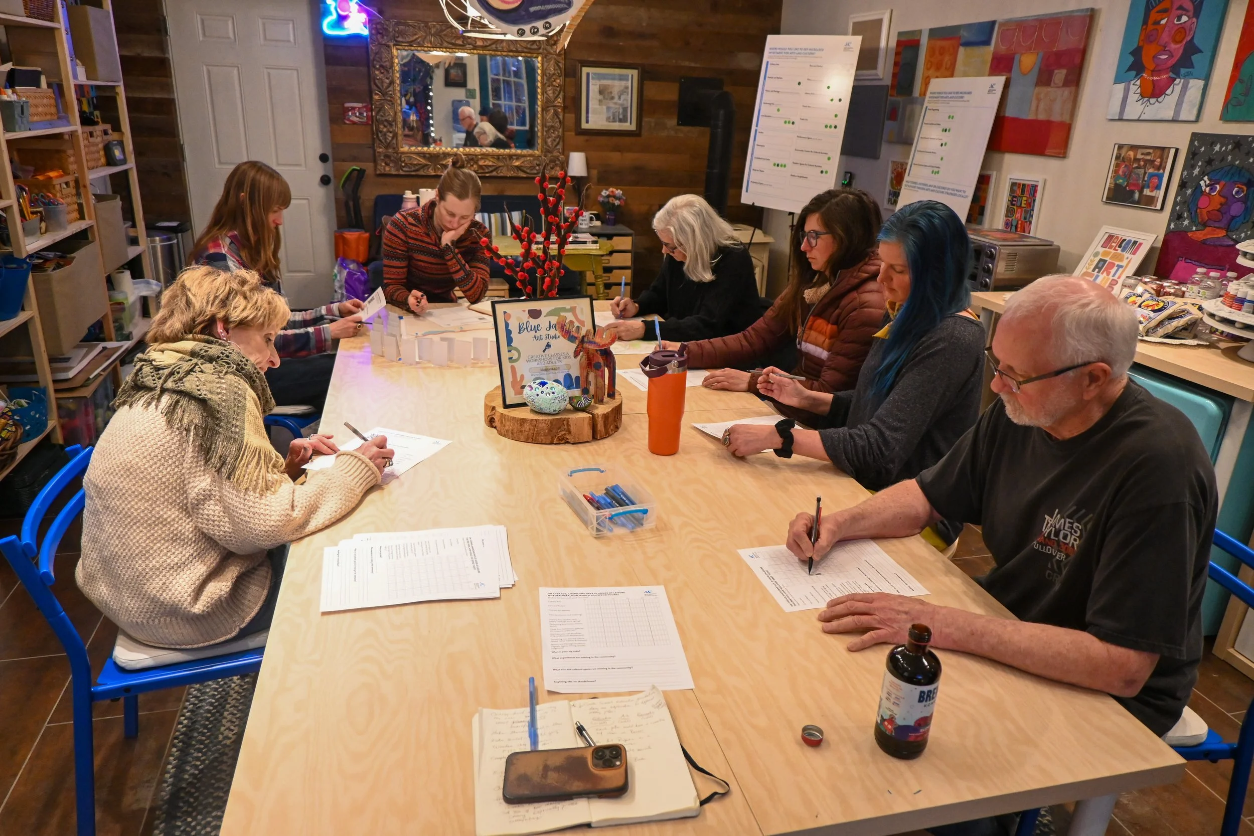 A group of eight people sitting around a large wooden table in a craft or art studio, engaged in writing or reading, with art supplies, papers, and notebooks on the table. The room has colorful art on the walls, shelves with supplies, and a mirror on