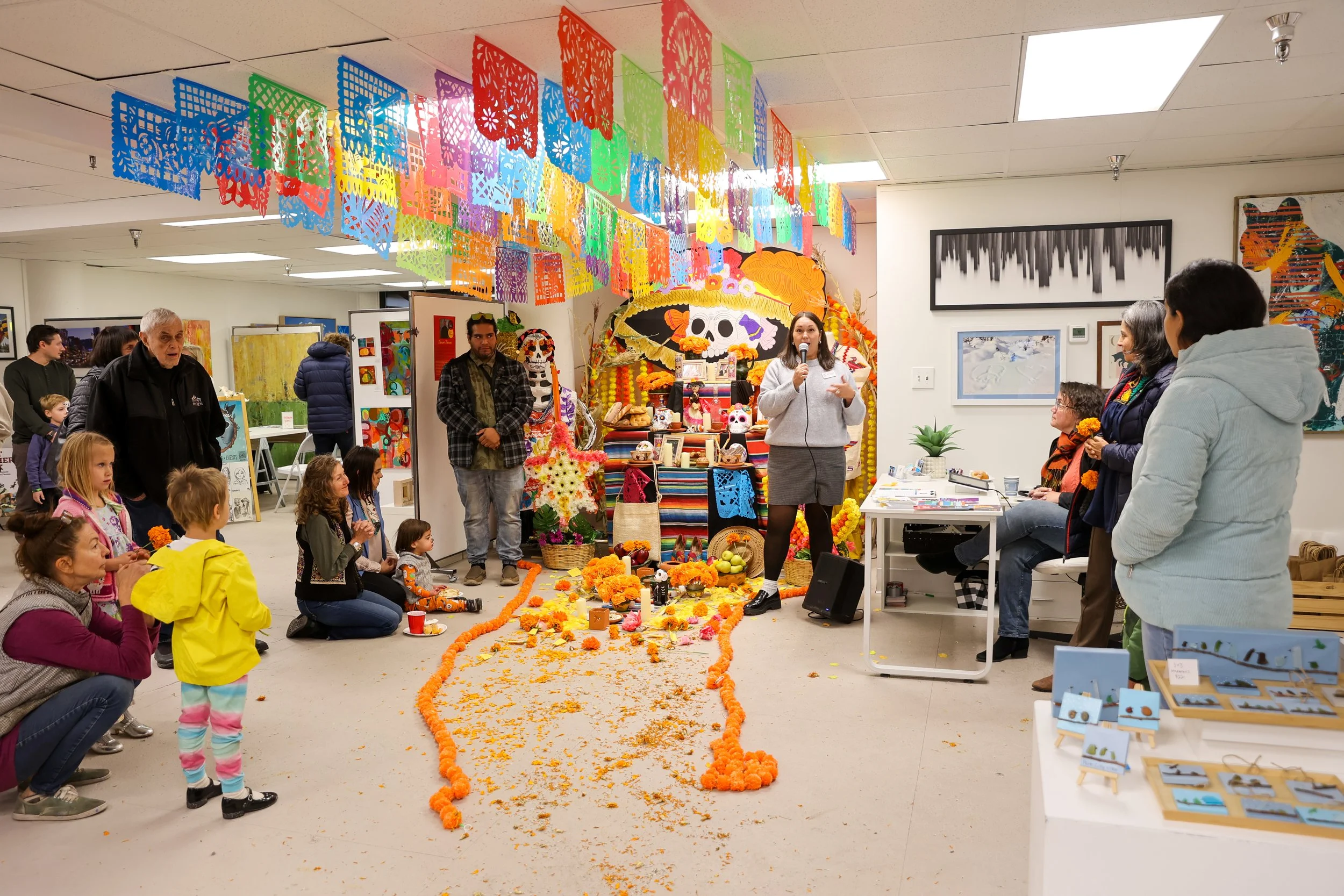 As pessoas reuniram-se numa galeria de arte durante uma celebração, com um altar decorado com malmequeres, motivos de caveiras e bandeiras de papel picado, todos indicativos do Dia dos Mortos.