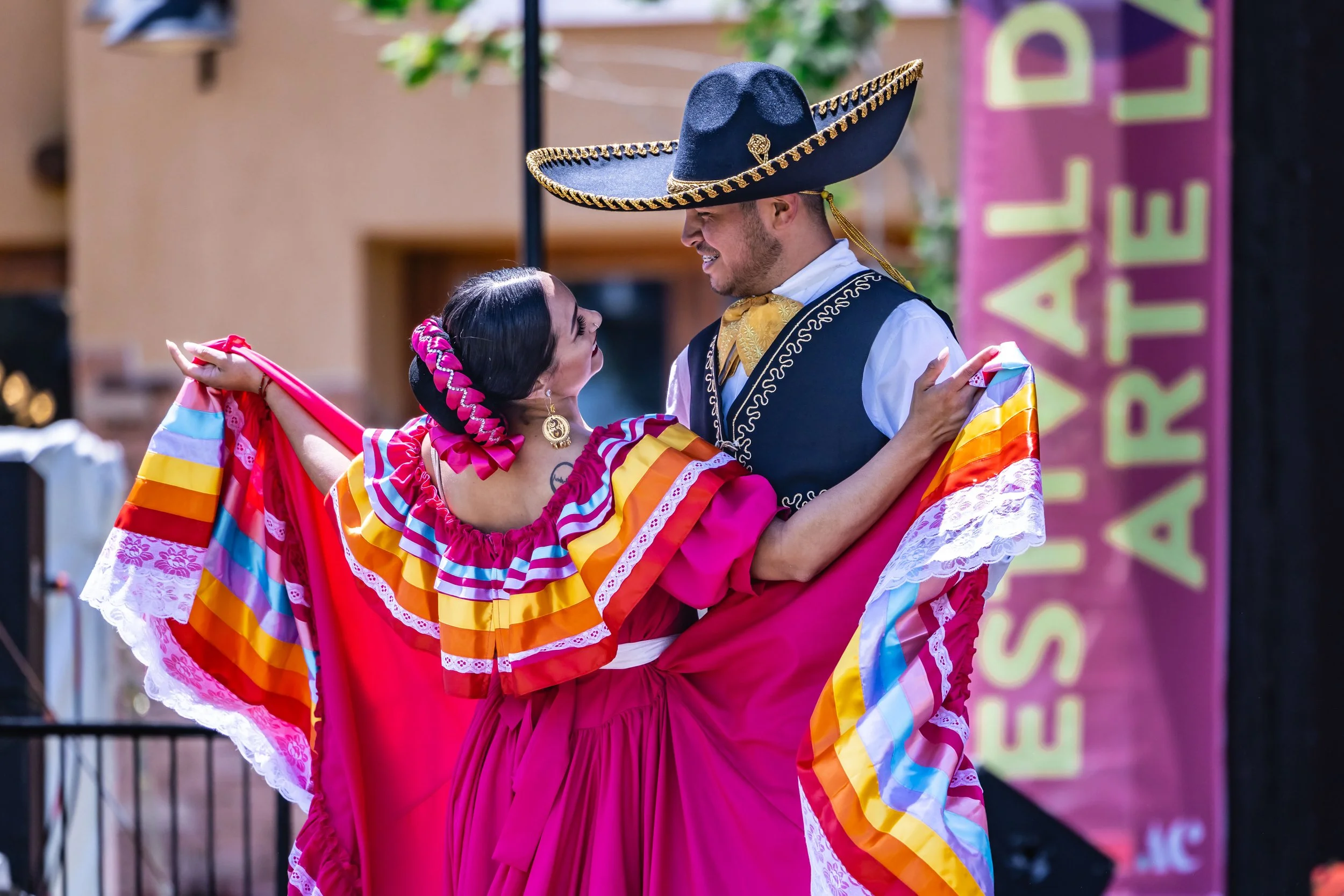 Um casal dançando com roupas tradicionais mexicanas coloridas, com um evento ao ar livre ao fundo e uma faixa com os dizeres «Festival de Arte Latino».