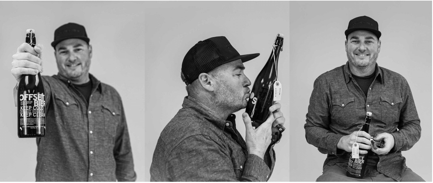 Three men in casual clothing and caps holding bottles of beer, with one man kissing his bottle, against a plain background.