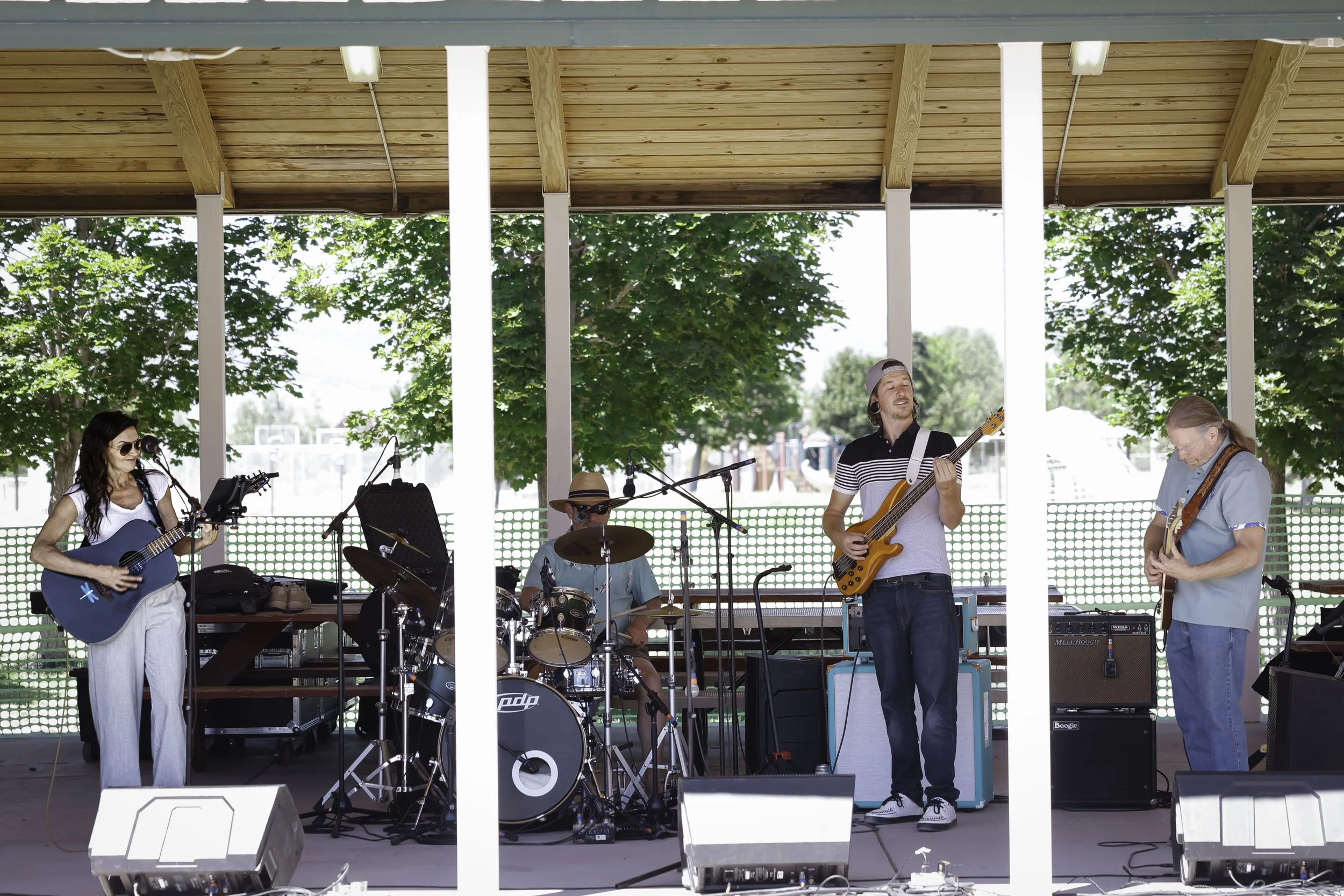 A four-piece band playing on an outdoor stage with trees in the background. The band includes a female guitarist, a male drummer wearing a hat, a male bassist, and a male guitarist. The stage has a wooden ceiling and is set up with microphones and amplifiers.
