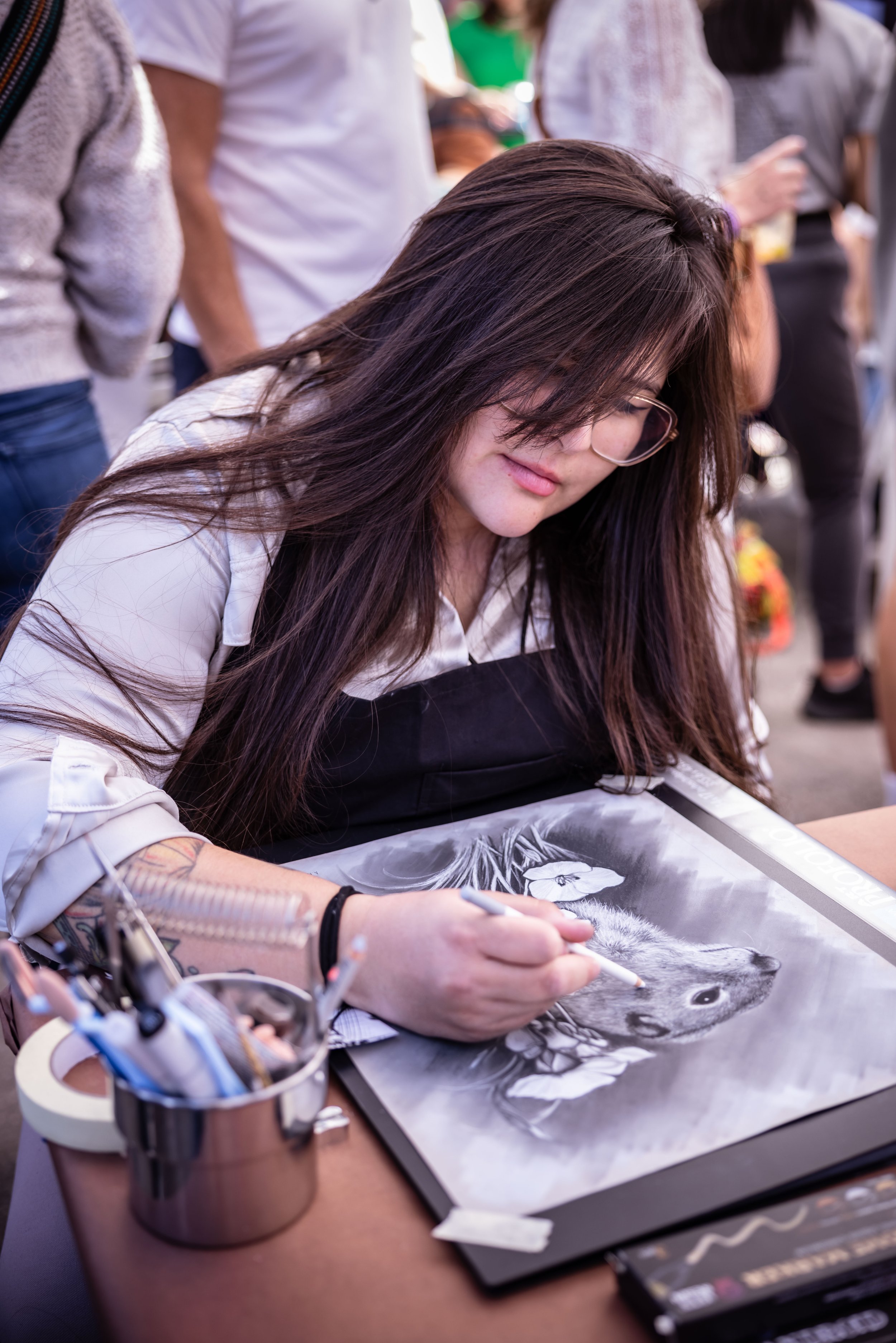 A woman with long dark hair and glasses drawing a highly detailed black and white artistic portrait of a wolf on paper at a table, with art supplies nearby at an outdoor event.