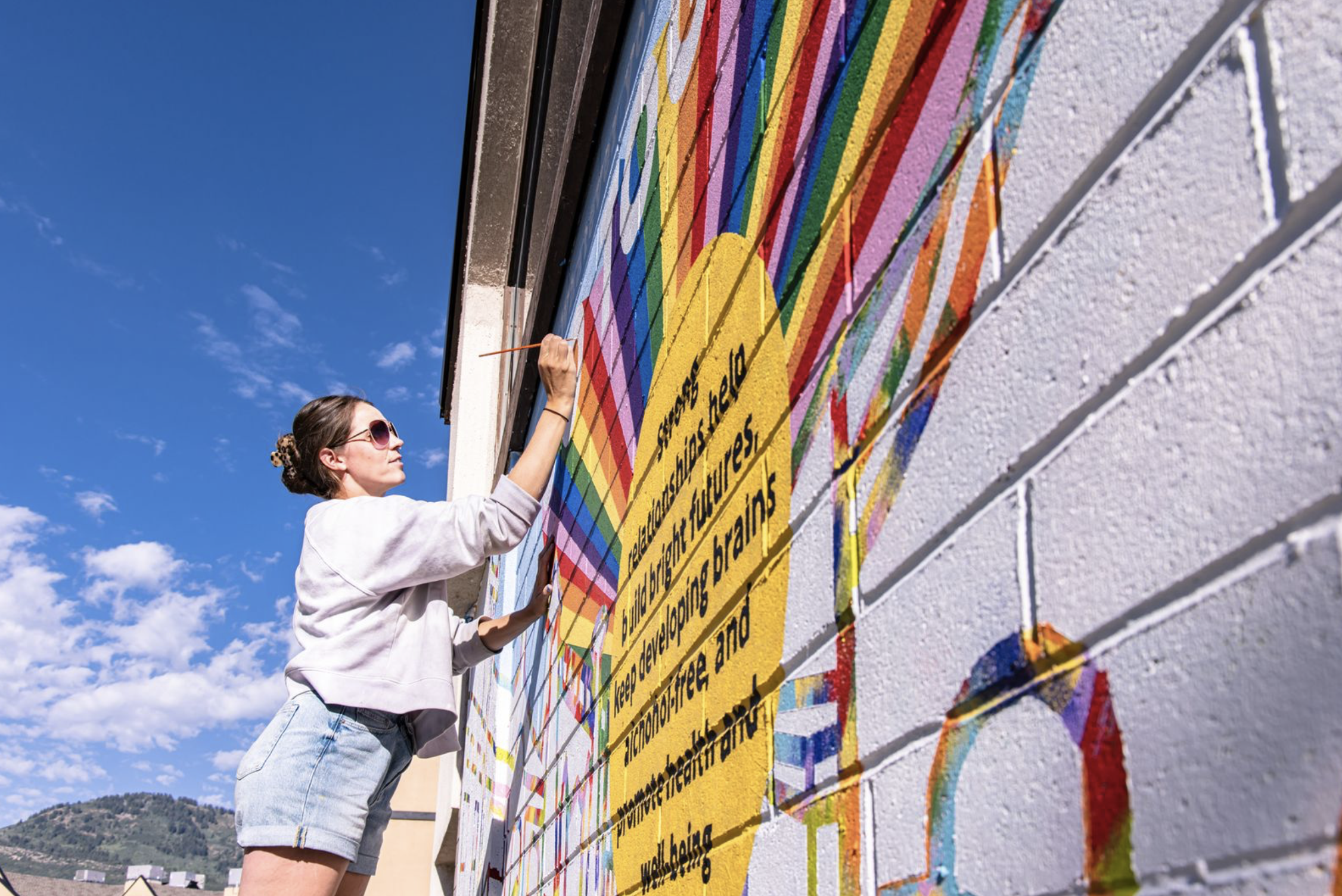 Woman painting a colorful mural on a brick wall outdoors, under a blue sky with some clouds.