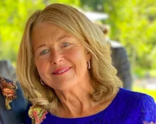 A middle-aged woman with blonde hair and blue eyes, smiling softly, outdoors with green trees in the background, wearing a blue top.