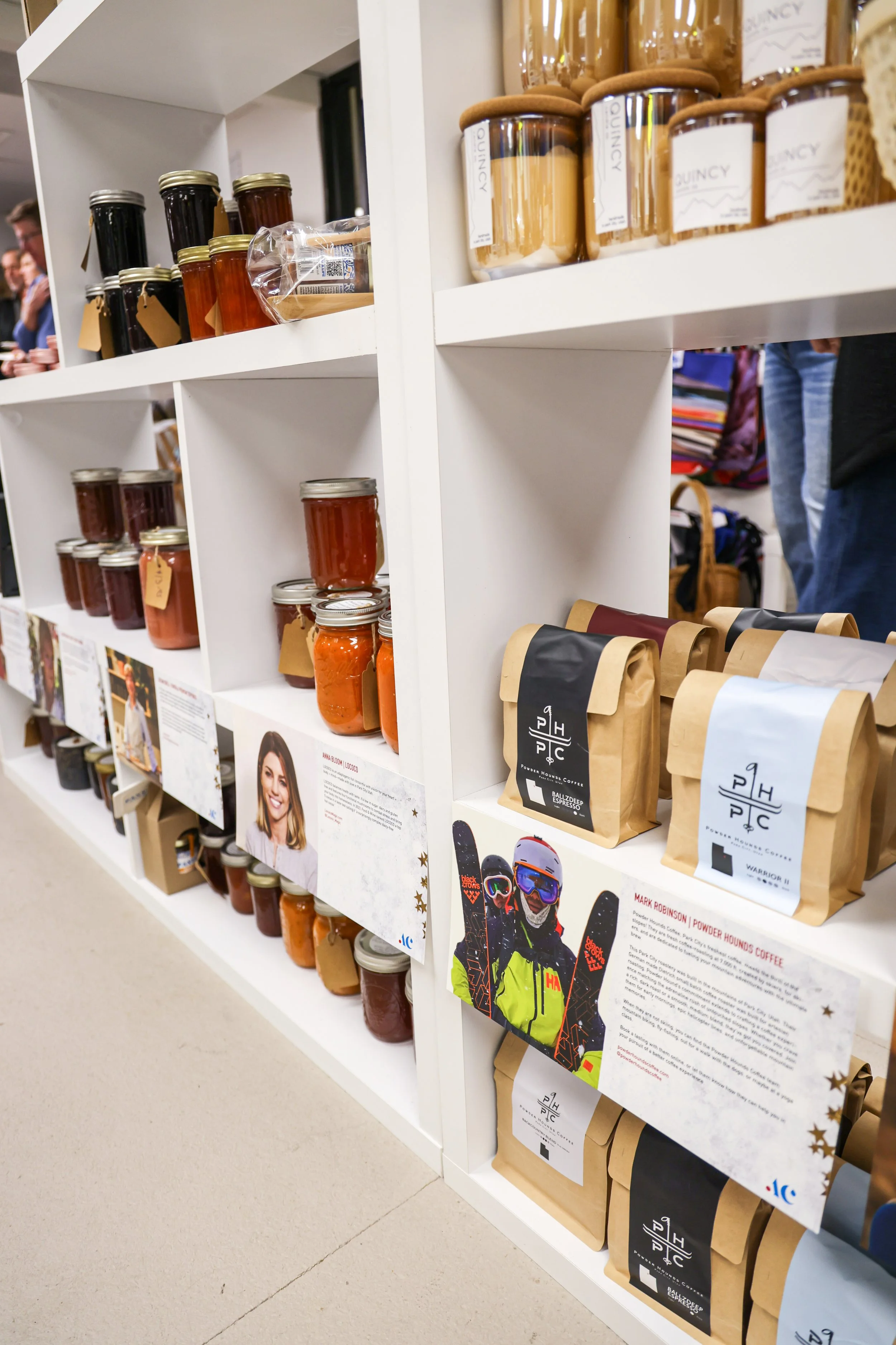 Shelves with jars of preserves and bags of coffee, along with informational cards and promotional photos, in a retail store.