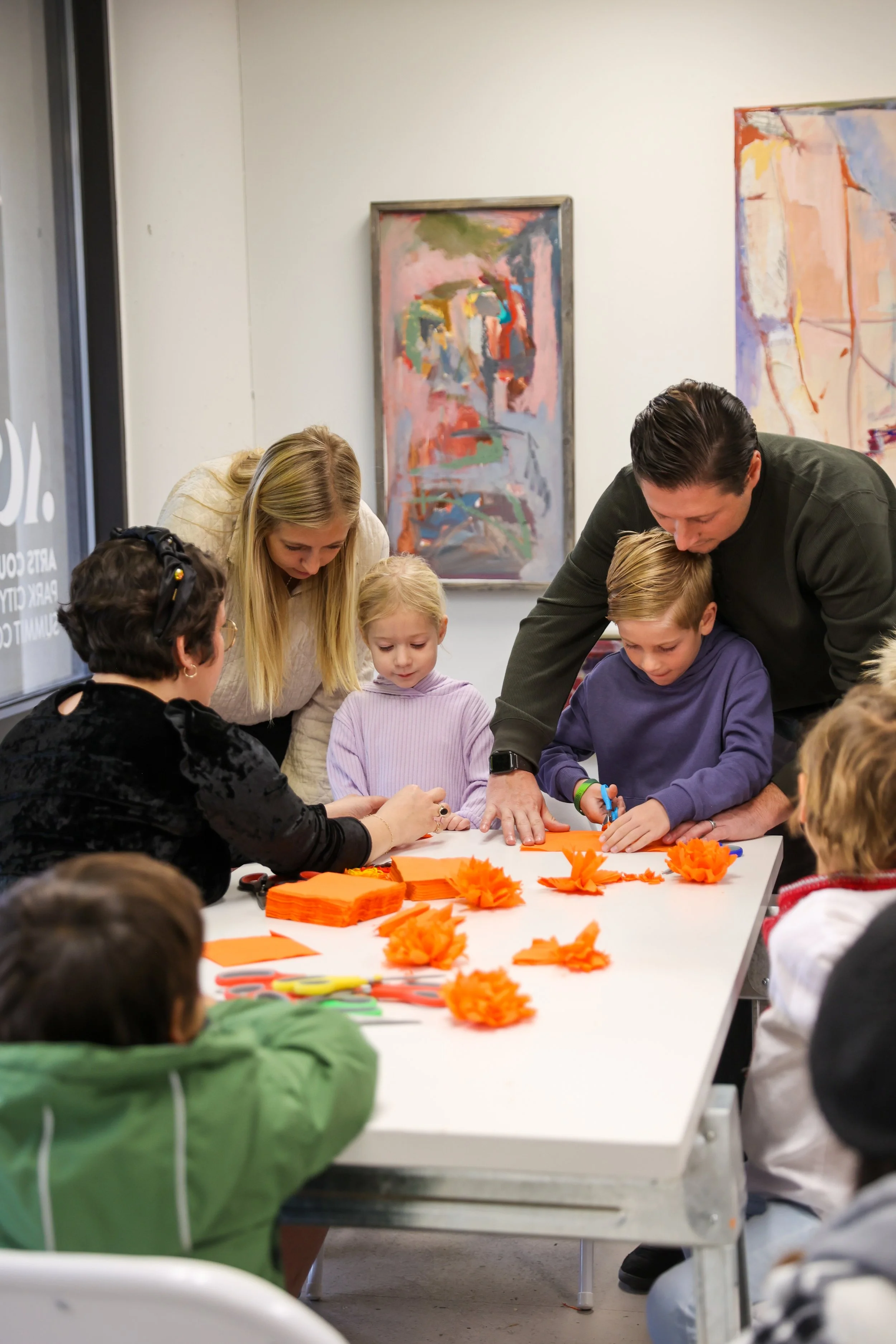 Adultos e crianças reuniram-se em torno de uma mesa para fazer flores de papel cor de laranja.