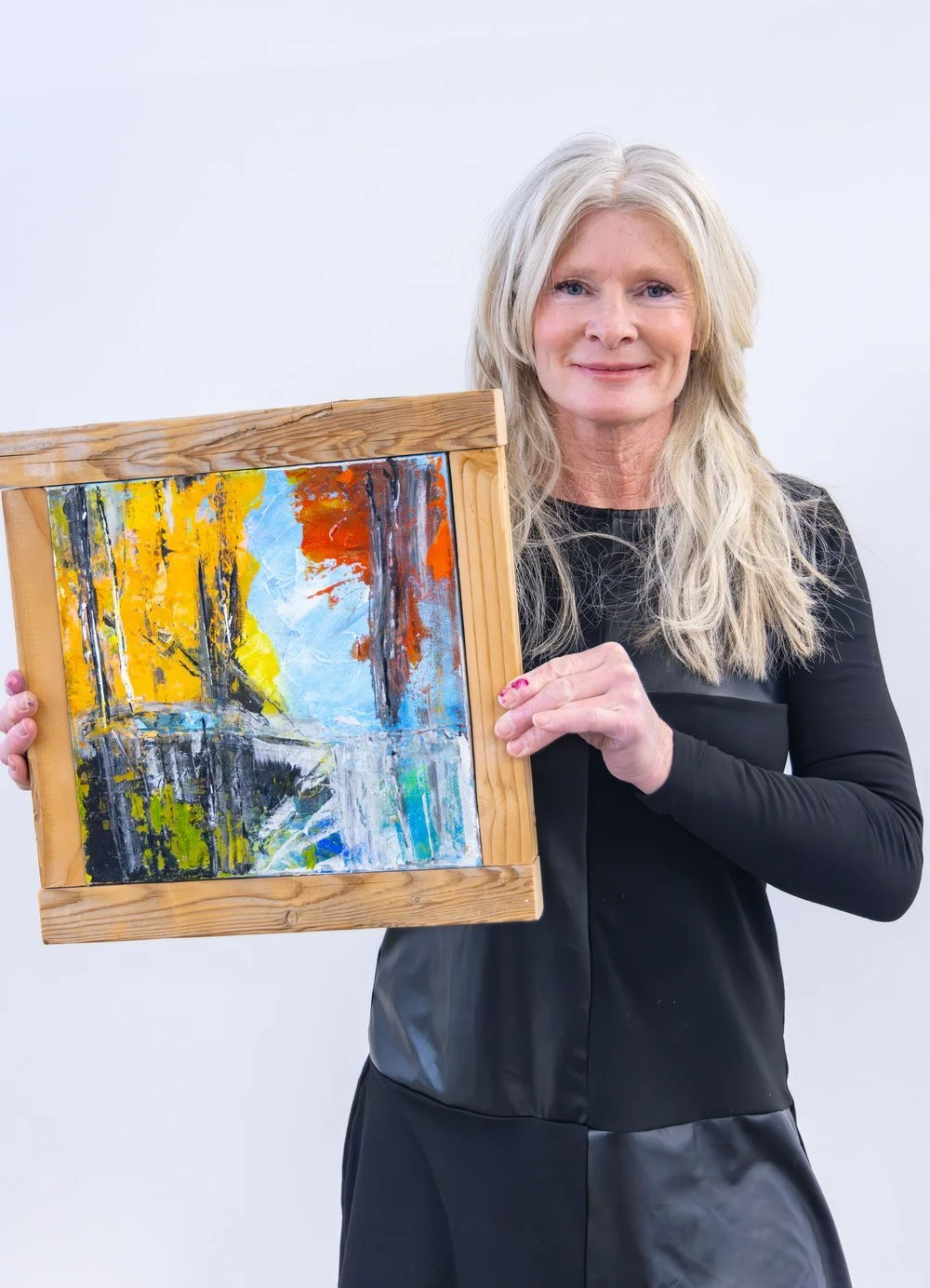 A woman with long gray hair holding a colorful abstract landscape painting in a wooden frame.