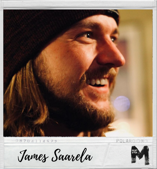 Close-up of a smiling man with long hair, beard, and a black beanie, holding a Polaroid photo with the name James Saarela written on it.