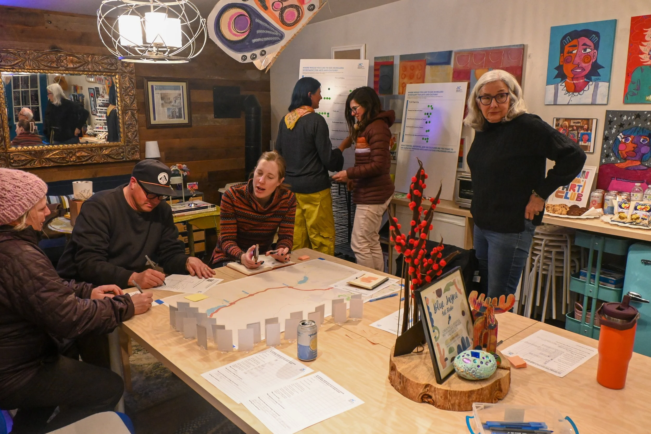 A group of people gathered around a table with a large map, engaging in a discussion and taking notes, in a warmly decorated room with artwork on the walls.