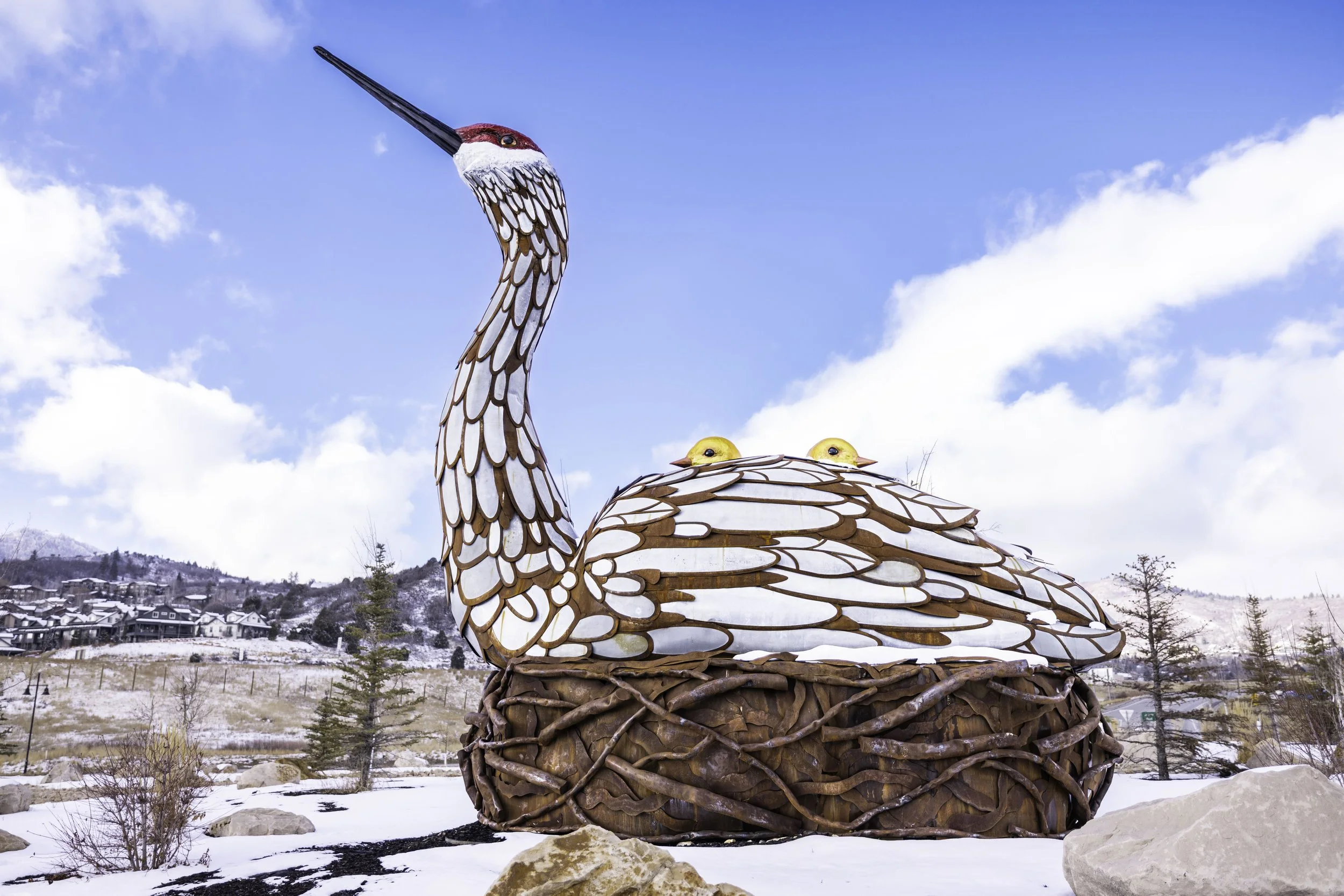 Large bird sculpture resembling a crane with a nest containing hatchlings, set outdoors in a snowy landscape with trees and houses in the background.