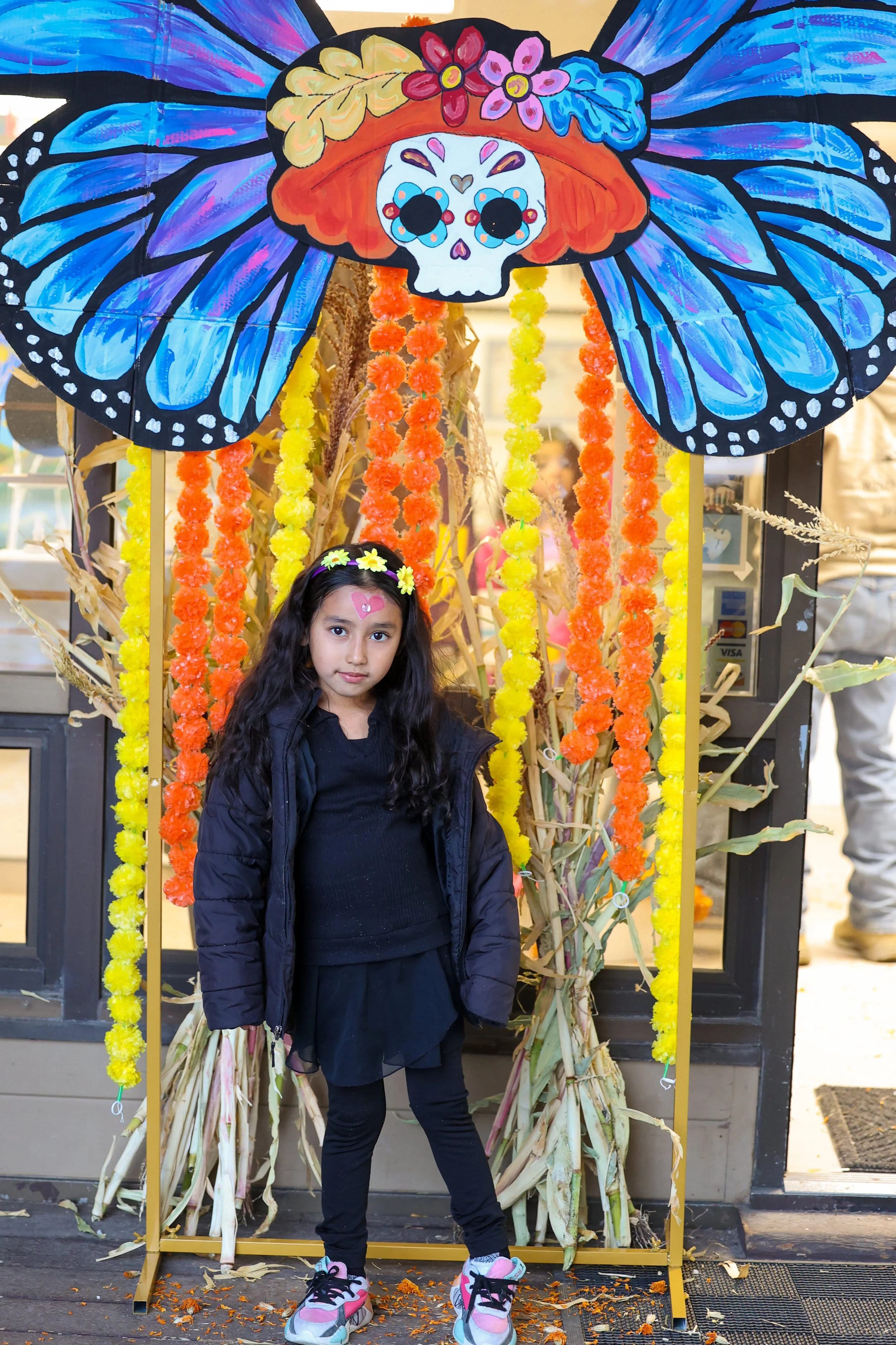 Jovem rapariga com cabelo comprido e escuro em pé em frente a um espantalho de abóbora decorado com guirlandas coloridas de malmequeres, uma borboleta pintada de azul e uma caveira de açúcar no topo, durante o festival de outono.