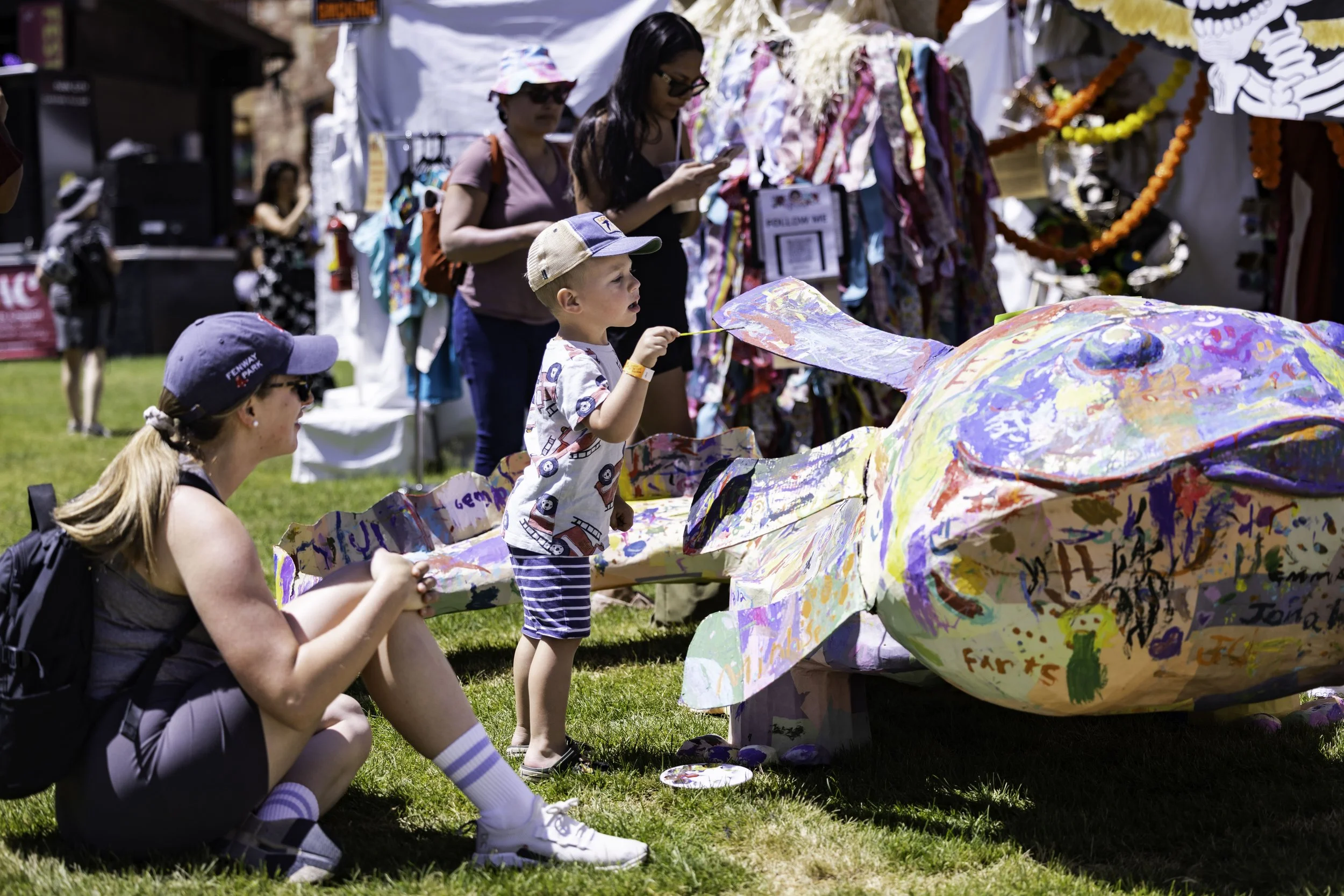 A young boy and a woman, possibly his mother, drawing and painting on a large, colorful, paper mache or cardboard sculpture at an outdoor art festival or fair on a sunny day. The woman is crouching while the boy is standing and using a paintbrush.