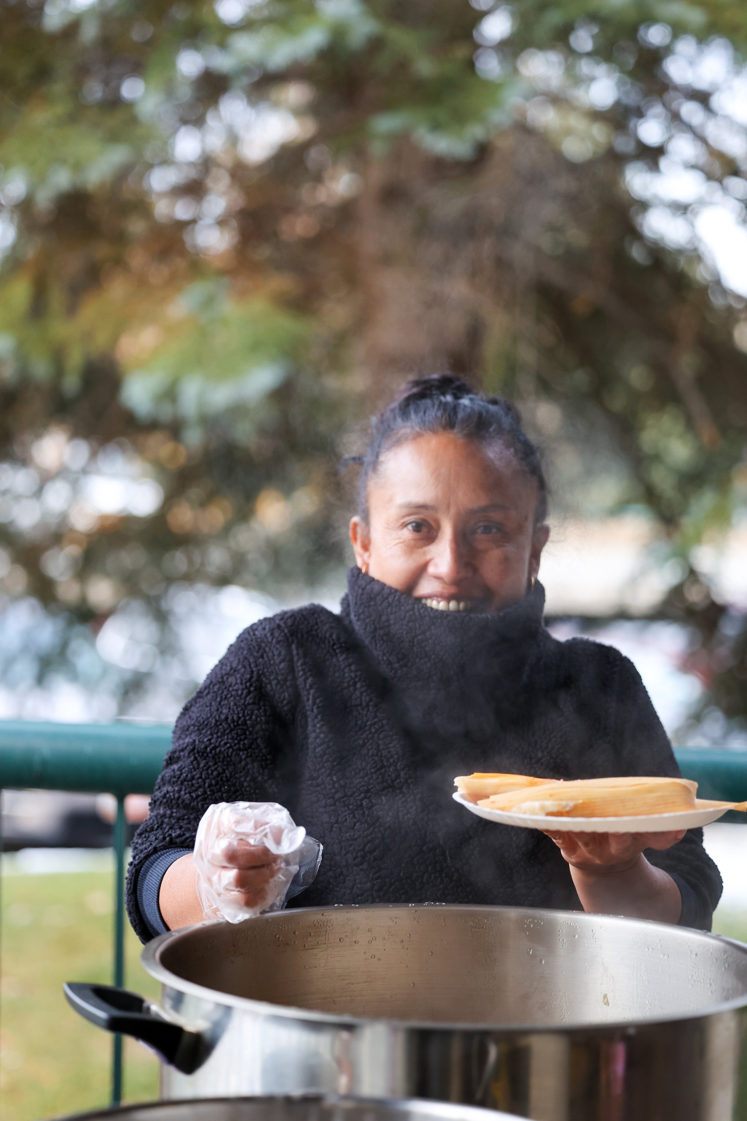 Mulher a servir comida quente de uma panela grande ao ar livre, sorrindo, com árvores verdes ao fundo.