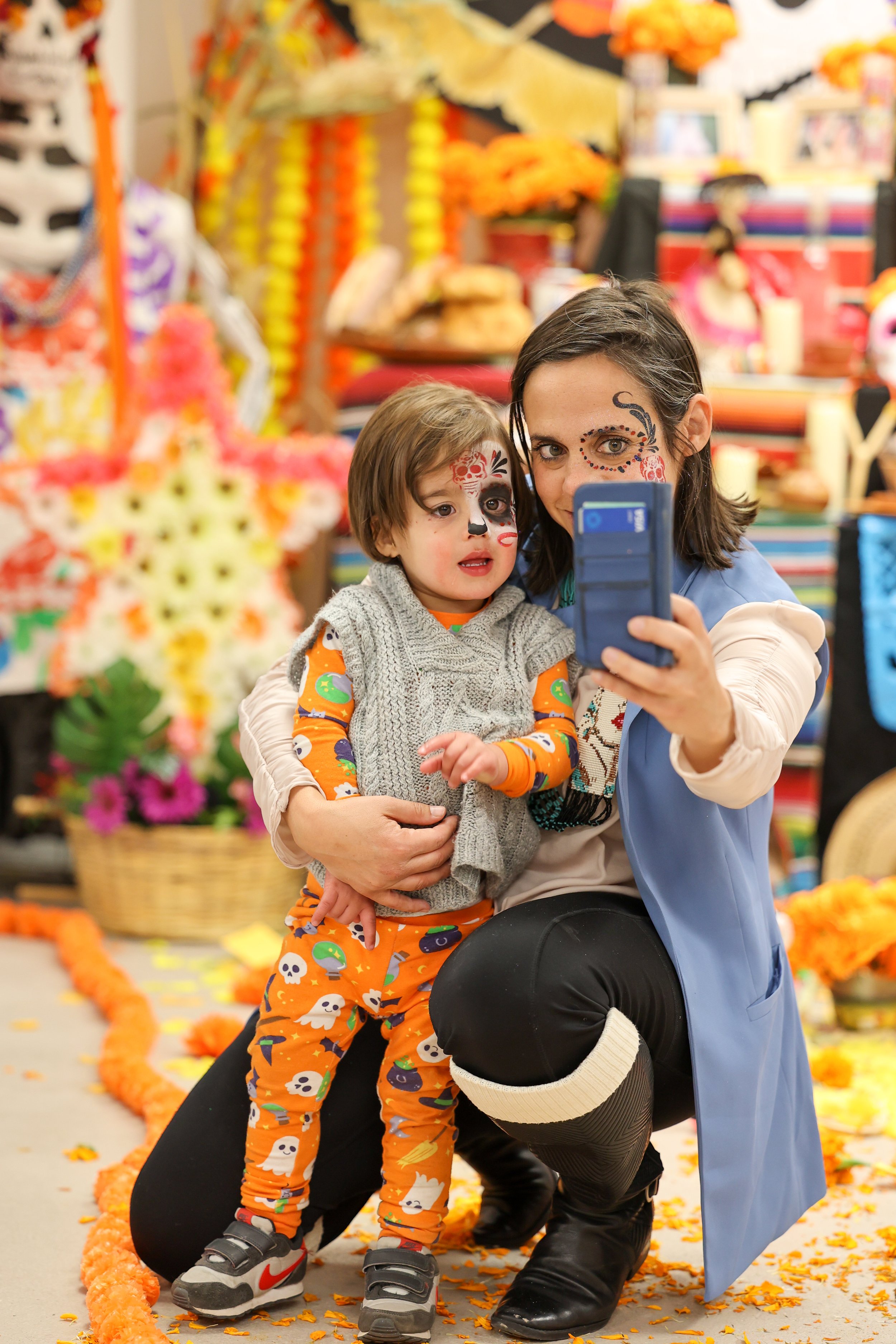 Uma mulher e um menino com pinturas faciais, tirando uma selfie numa celebração ou festival com decorações de Halloween e Dia de los Muertos, incluindo malmequeres, velas e faixas coloridas.