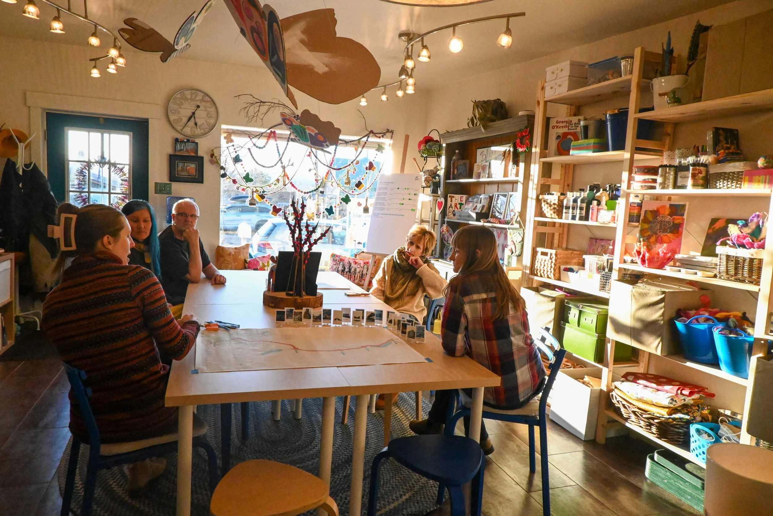 Five people sitting around a rectangular table in a cozy, decorated room with shelves full of art supplies, paintings, and craft projects, engaging in a creative activity.