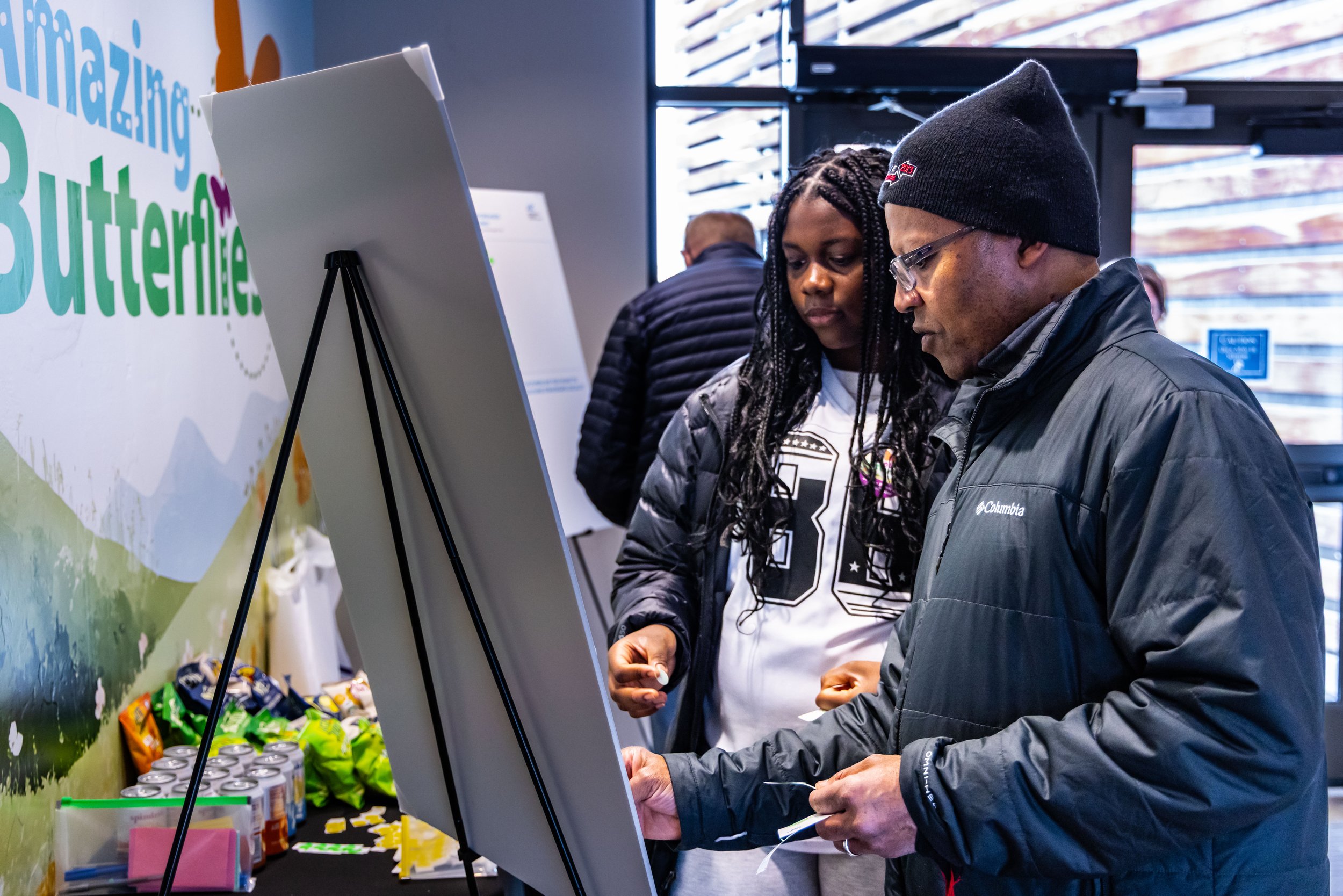 A man and a woman looking at a display board at an indoor event, with snacks and soda cans on a table nearby.