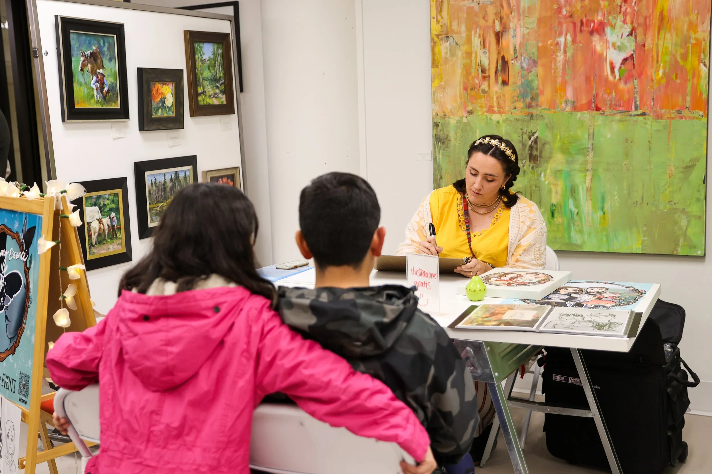 Um artista está sentado a uma mesa a autografar obras de arte para dois jovens visitantes numa galeria de arte. A galeria tem várias pinturas nas paredes, incluindo arte abstrata colorida atrás do artista e pinturas emolduradas de paisagens e retratos à sua esquerda. O visitante