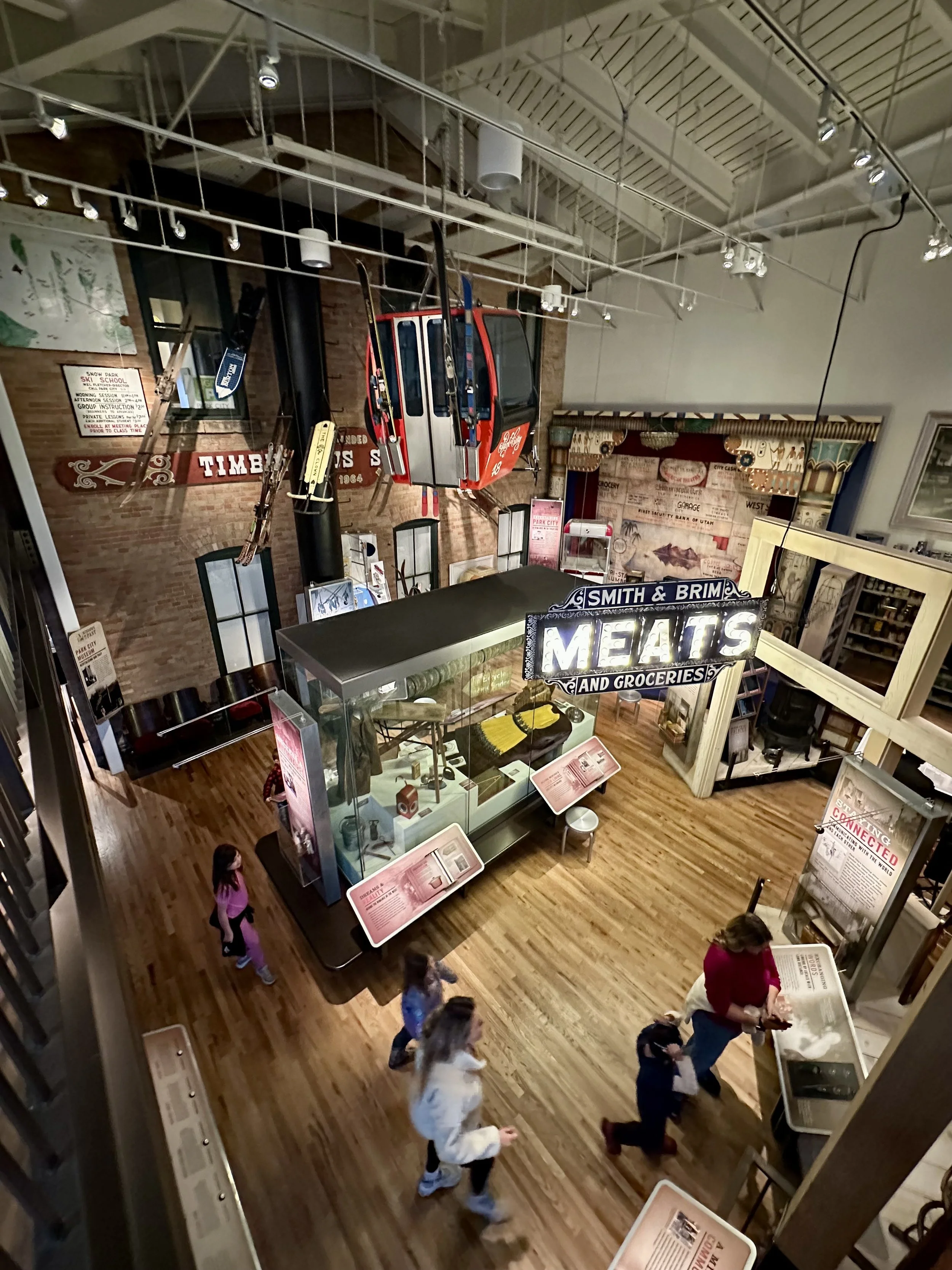 Una exposición en un museo interior con una telesilla suspendida de color rojo y gris y carteles antiguos. Hay vitrinas, carteles informativos y visitantes caminando sobre un suelo de madera.