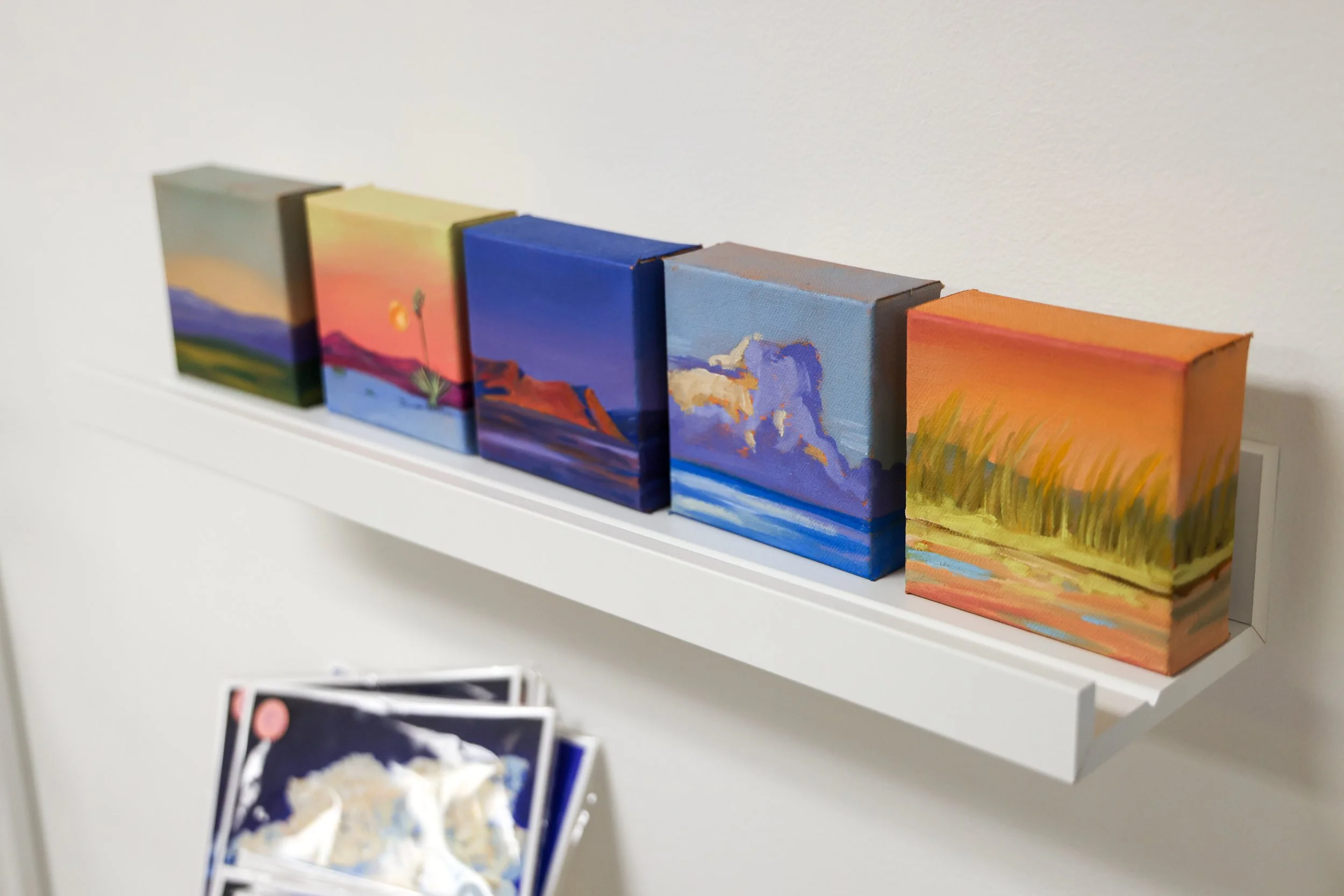 Six small landscape paintings displayed on a white shelf, depicting various sunset and mountain scenes in vibrant colors.