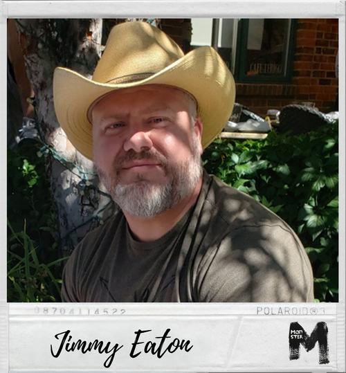 A man wearing a cowboy hat and a black shirt, sitting outdoors with greenery around and a brick building in the background.