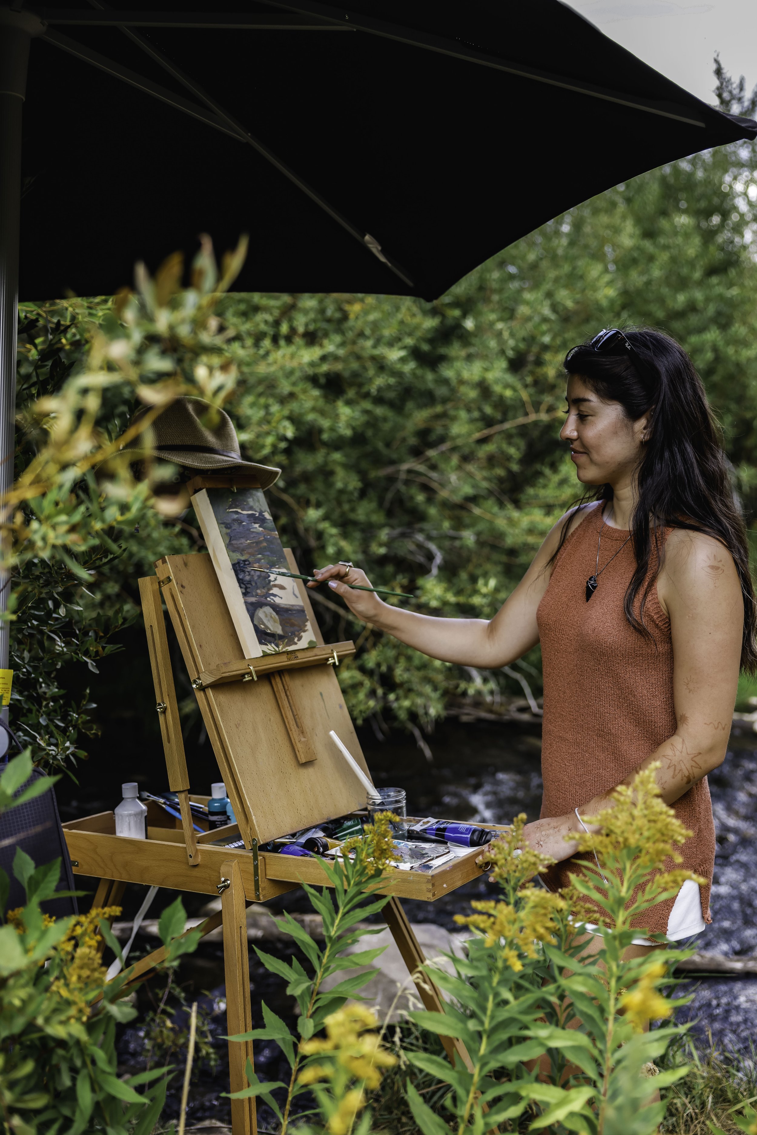 A woman painting outdoors under a black umbrella, surrounded by green foliage, with a wooden easel and art supplies.