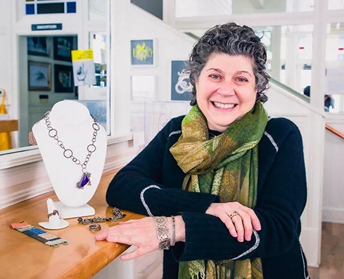 A woman with short curly gray hair smiling and sitting behind a display of jewelry in a store.