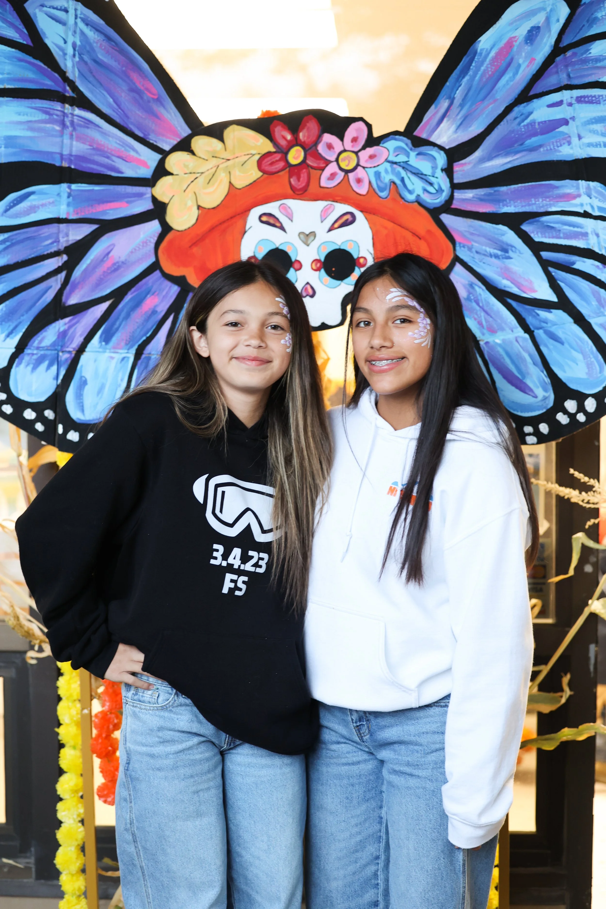 Duas meninas com pinturas faciais e roupas casuais em frente a uma grande pipa em forma de borboleta decorada com uma caveira de açúcar e flores.