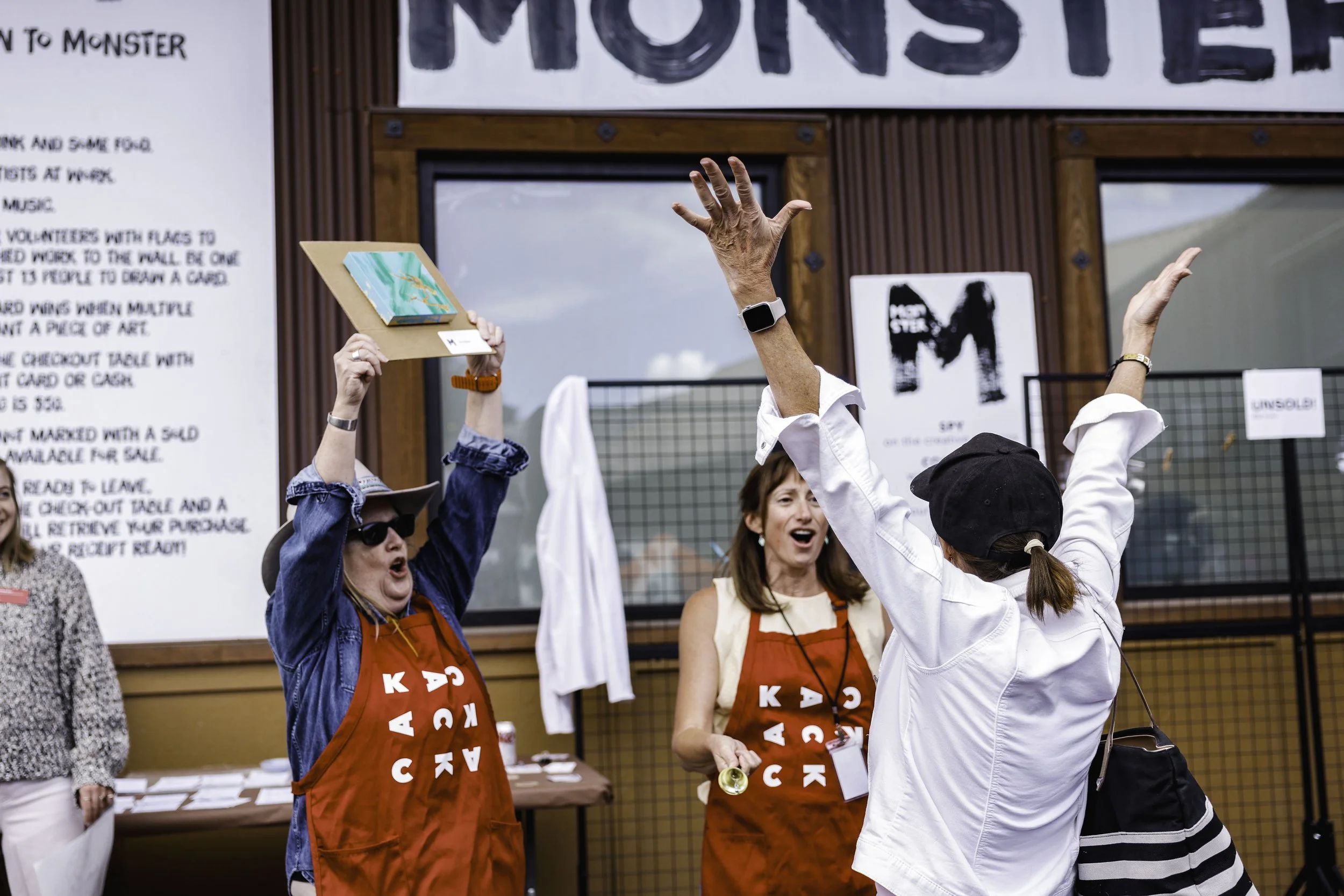 Two women wearing red aprons are celebrating outdoors, one holding a small painting and the other raising her hands, with a group of people and a sign in the background.