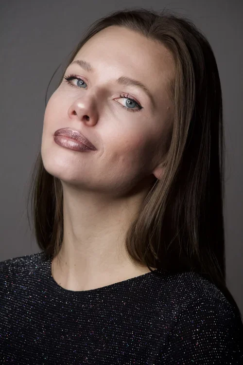 Close-up of a woman with long brown hair, light makeup, and a confident expression, wearing a black sparkly top against a gray background.
