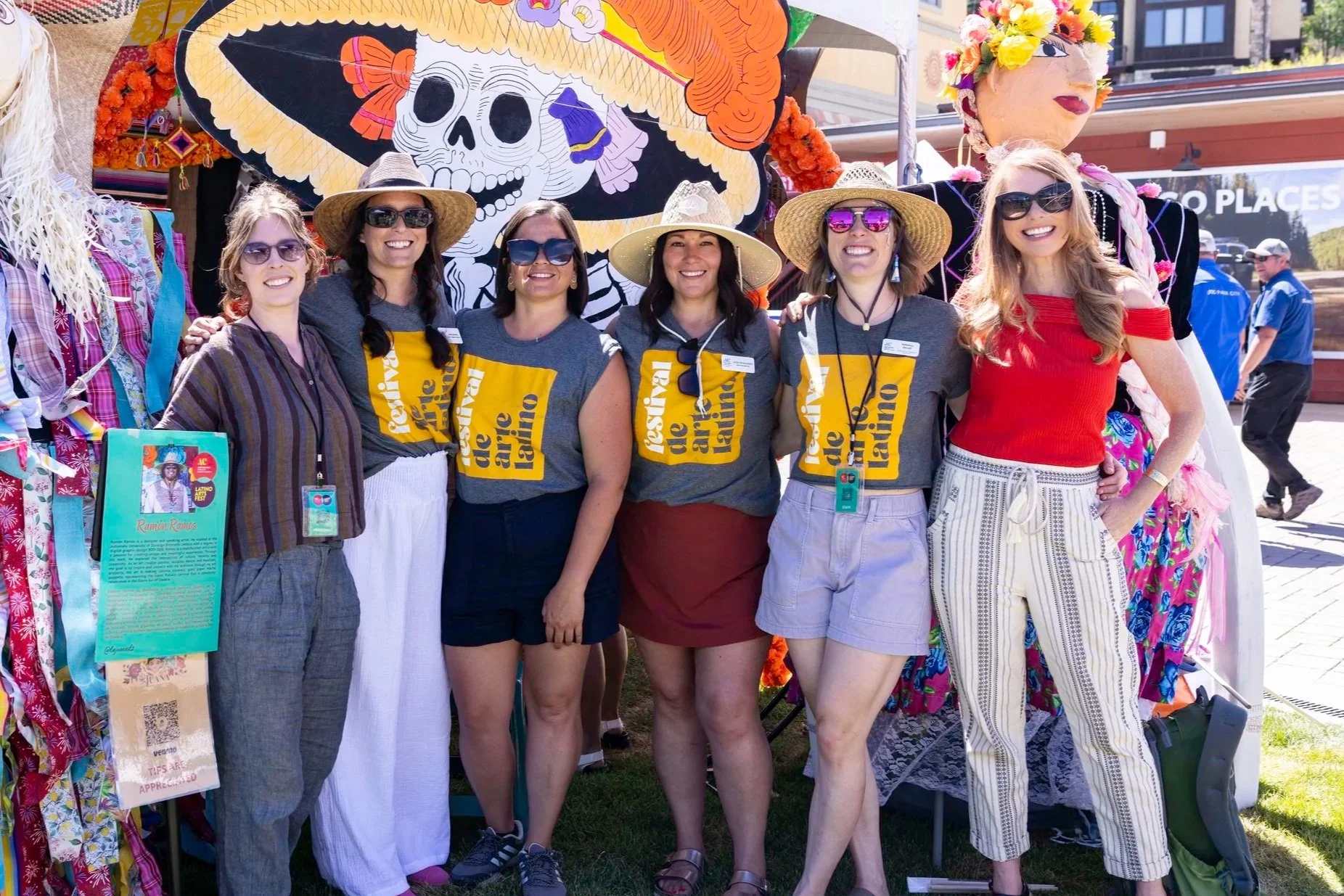 Group of six women standing together outdoors at a festival or event, wearing casual clothing and sunglasses, some with straw hats, smiling at the camera. Behind them are colorful decorations including a large skeleton figure with a hat, a floral headdress, and a vibrant art display.