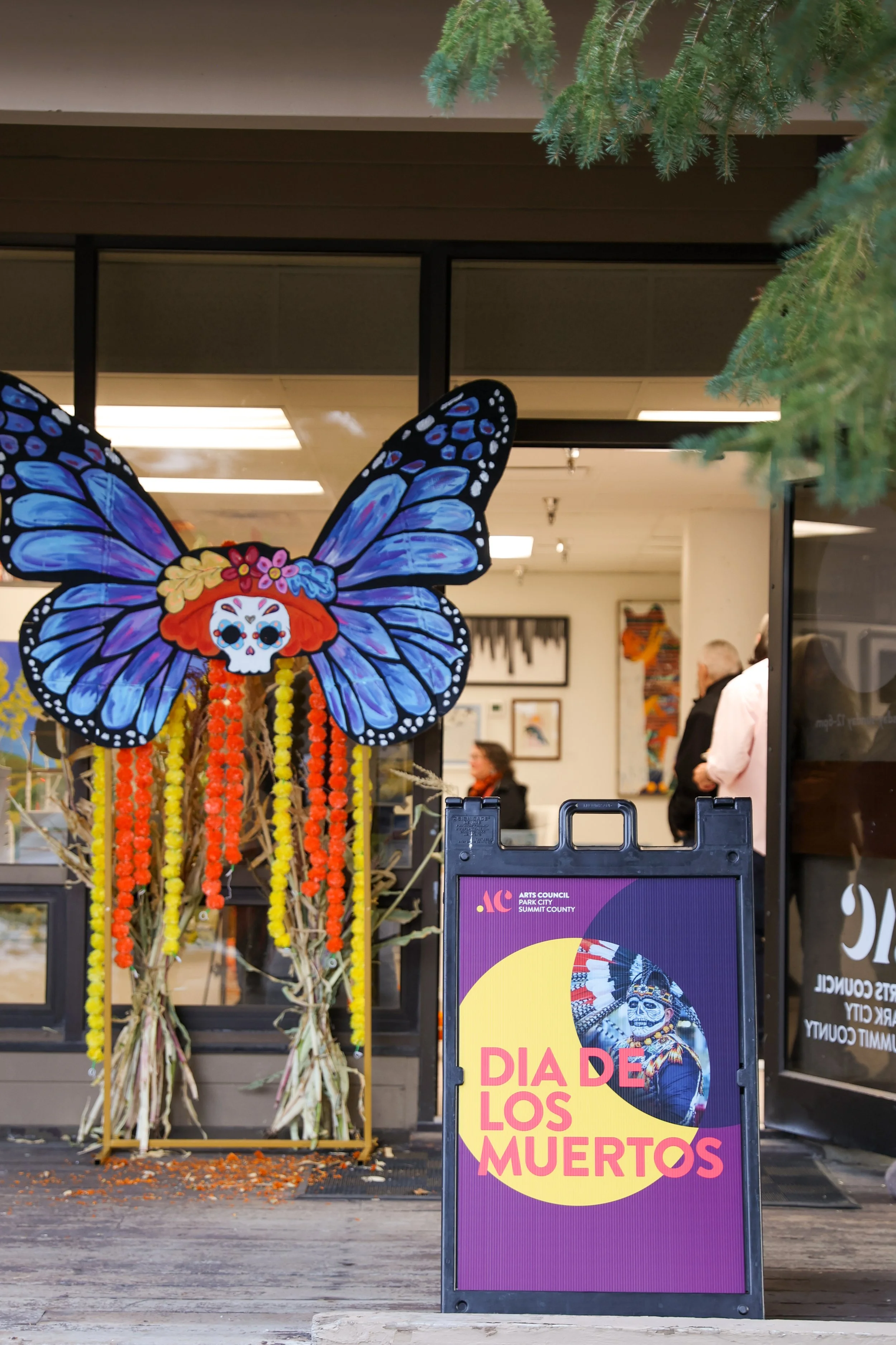 Exposição colorida do Dia dos Mortos com uma grande decoração em forma de borboleta e um cartaz promovendo a celebração do Dia dos Mortos.