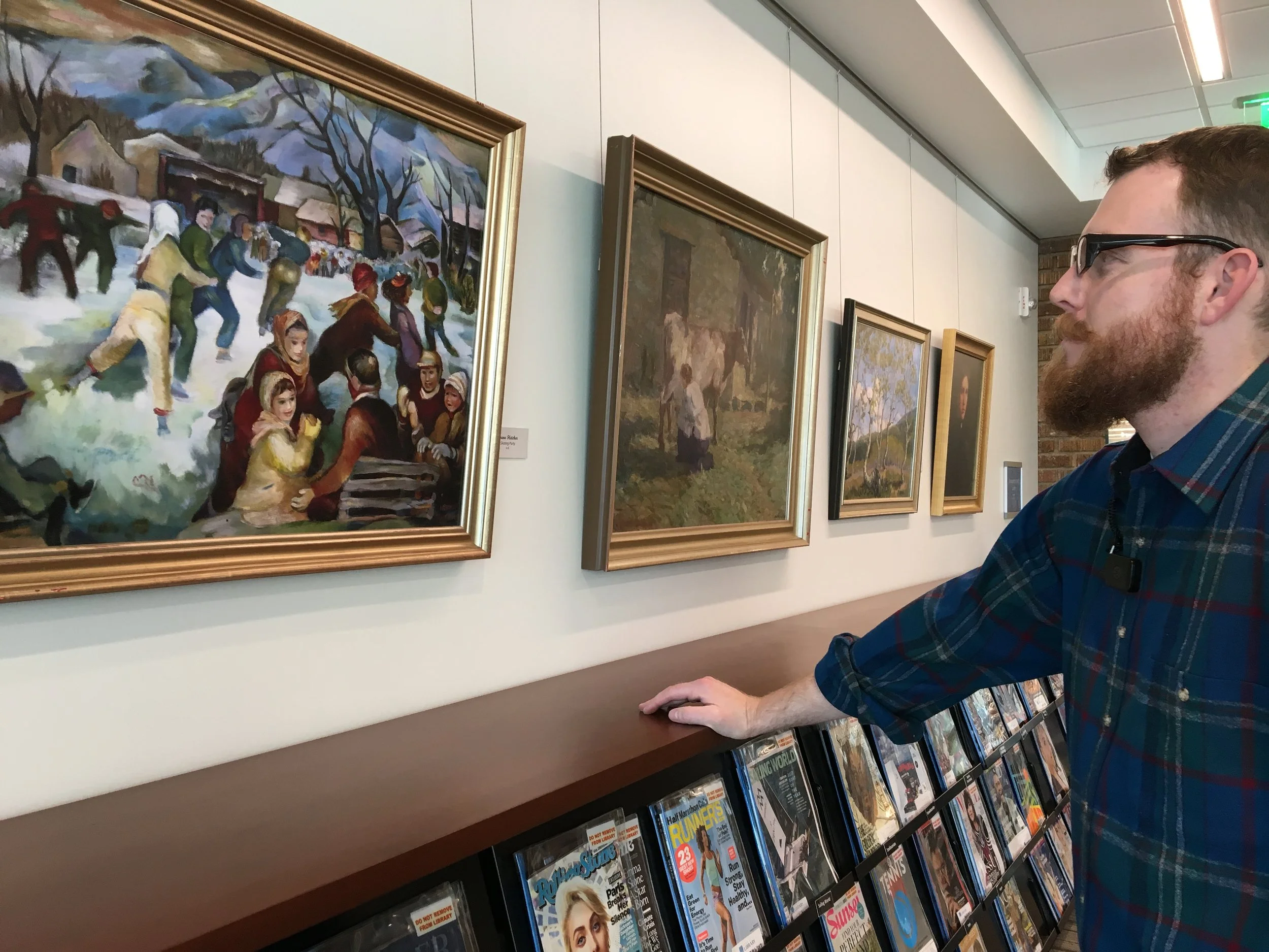 Un hombre con gafas, barba y camisa a cuadros está mirando cuadros en una galería de arte. Los cuadros representan escenas al aire libre y una escena invernal con gente jugando en la nieve.