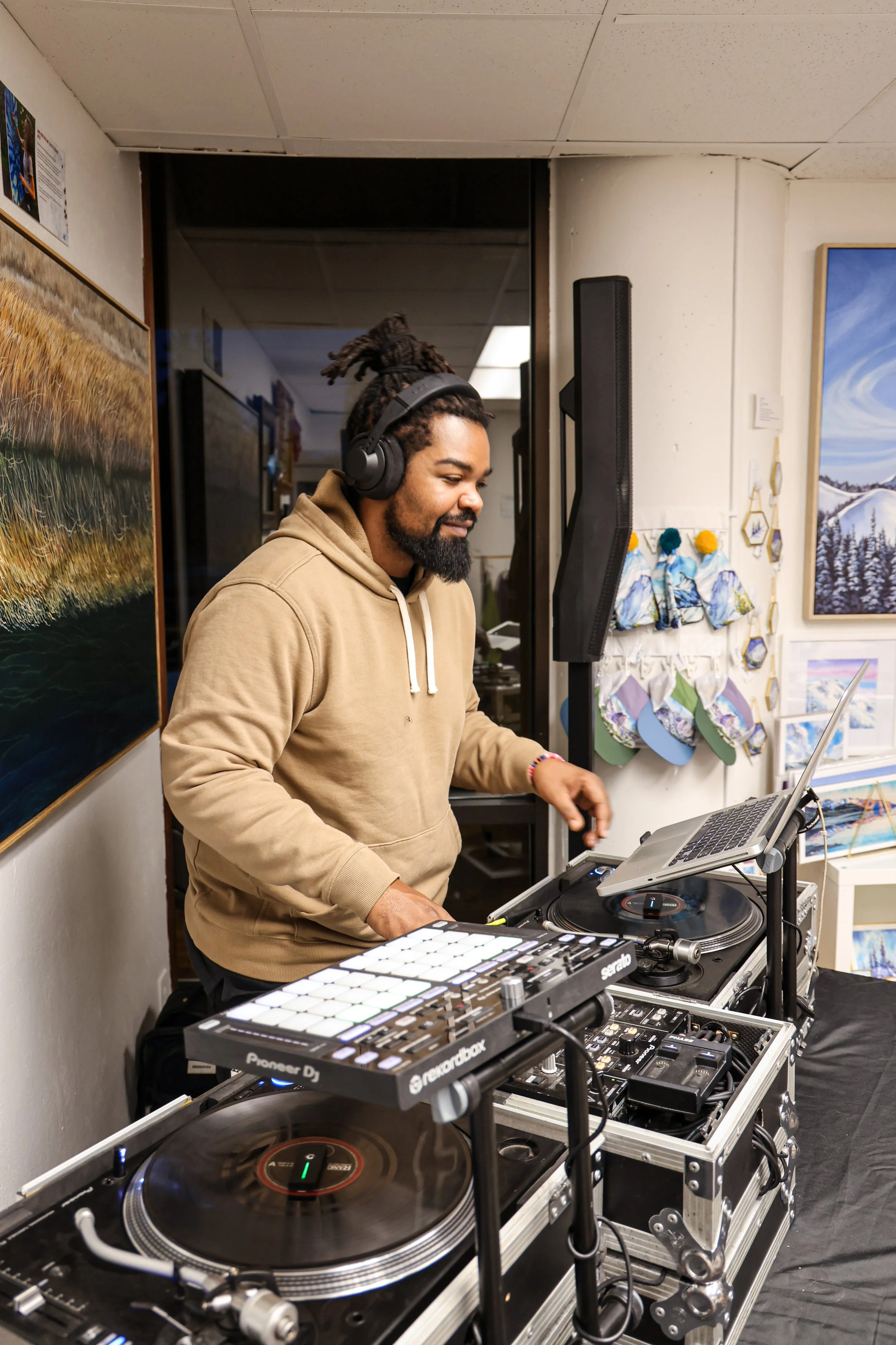 A man with dreadlocks and a beard wearing a tan hoodie, smiling while DJing with headphones on, set up with DJ equipment, a laptop, and a turntable in an art gallery with paintings and crafts on the wall.