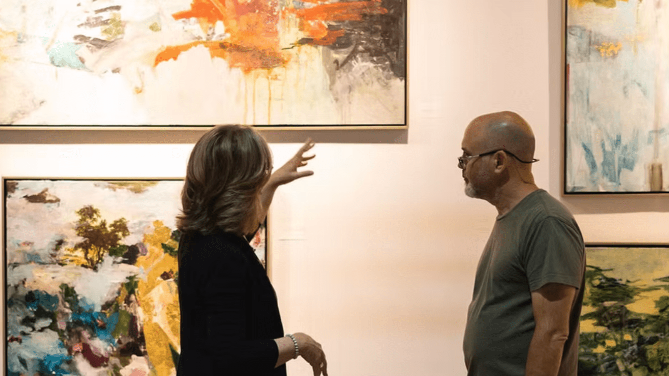 Two people, a woman with shoulder-length hair and a man with glasses and a beard, are engaged in conversation in an art gallery. The woman is gesturing towards a colorful abstract painting on the wall, which has orange, black, and white hues. The man is listening attentively.