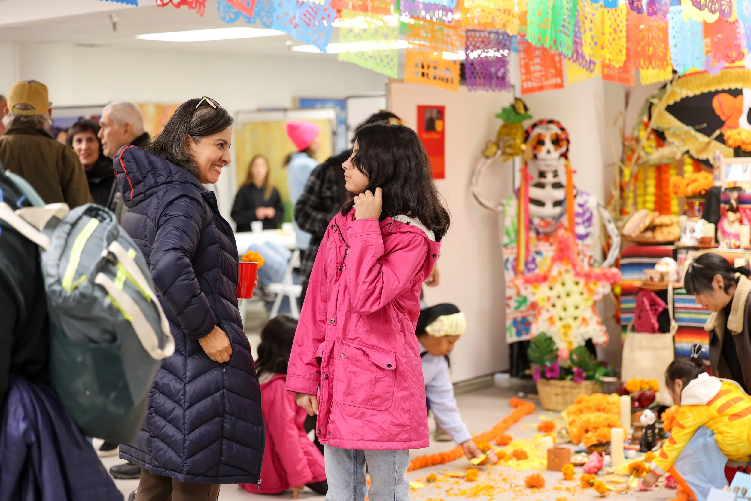 Duas mulheres conversando numa sala decorada com faixas coloridas de papel picado e enfeites festivos, com crianças e adultos ao fundo participando de uma celebração.