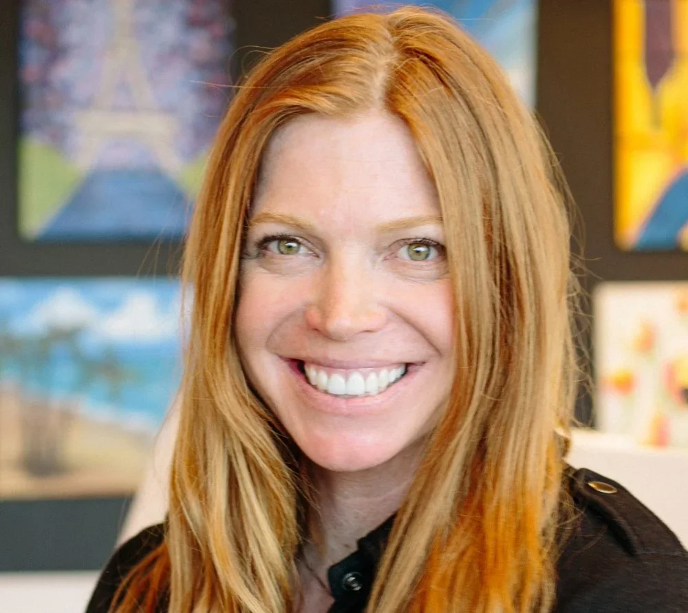 A woman with long red hair and a bright smile, wearing a black top, in front of colorful artwork.