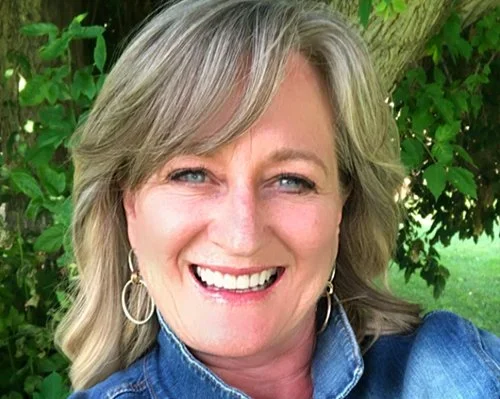 A woman with blonde hair smiling outdoors, wearing hoop earrings and a denim shirt, with green trees and grass in the background.