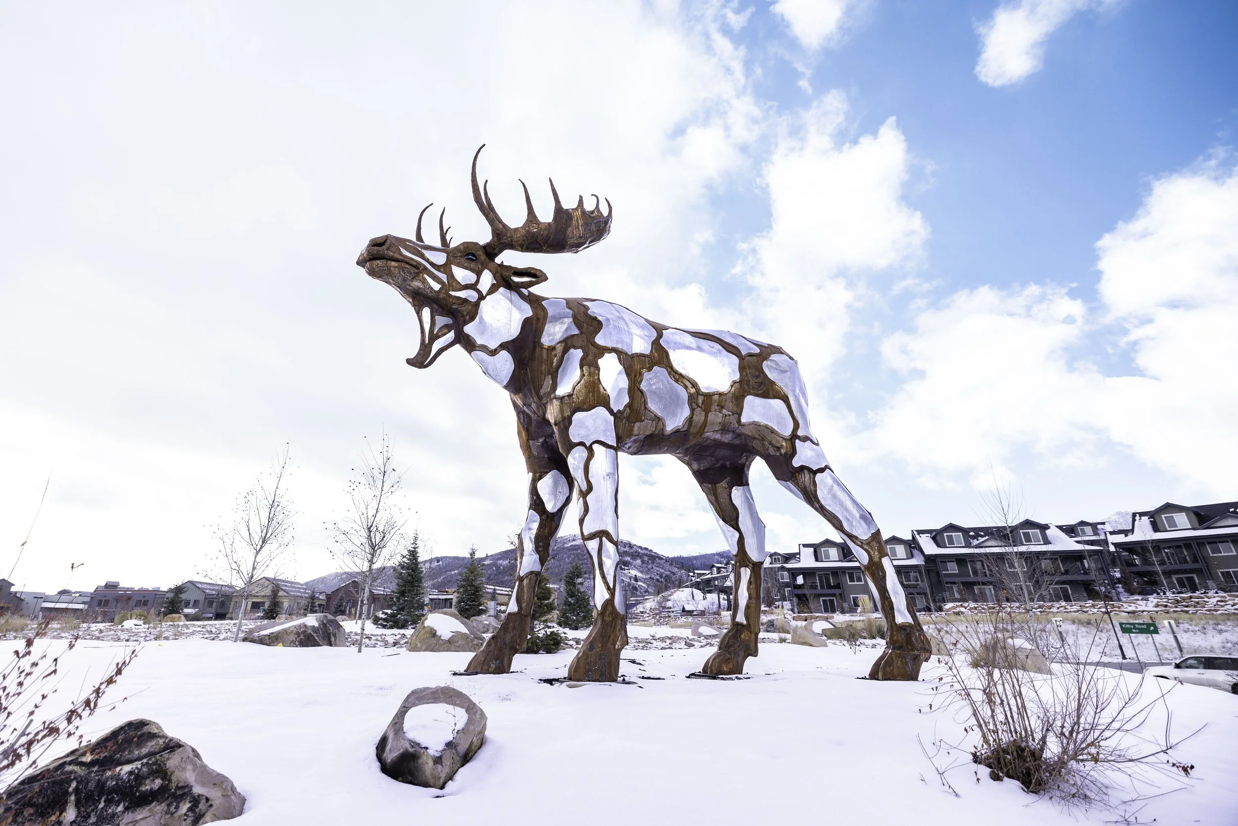 Large wooden sculpture of a moose, set outdoors in a snowy landscape under a partly cloudy sky, with residential buildings and trees in the background.