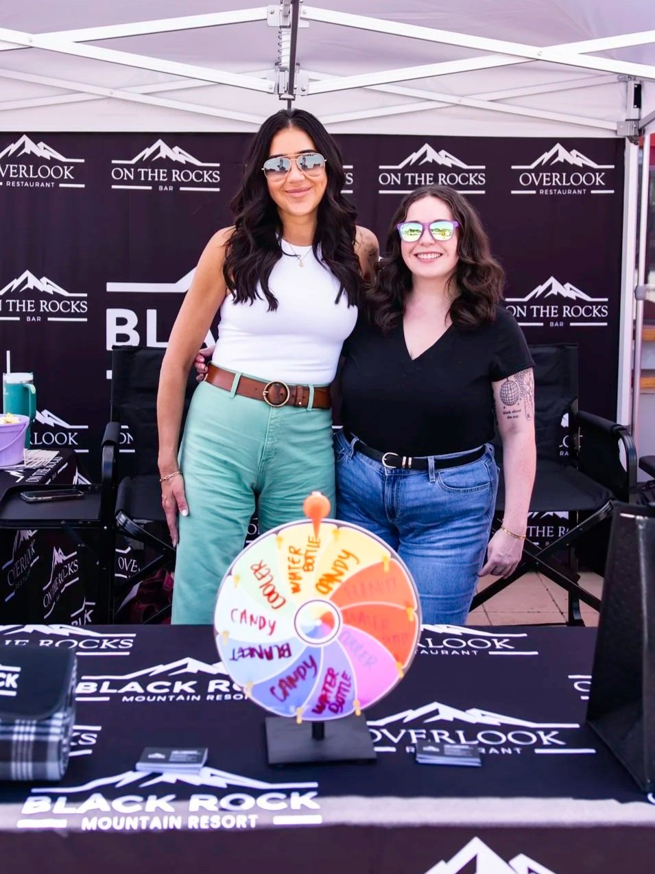 Two women standing behind a table with a spinning wheel game, at an event at Black Rock Mountain Resort, with a backdrop displaying logos for Overlook Restaurant and On The Rocks Bar.
